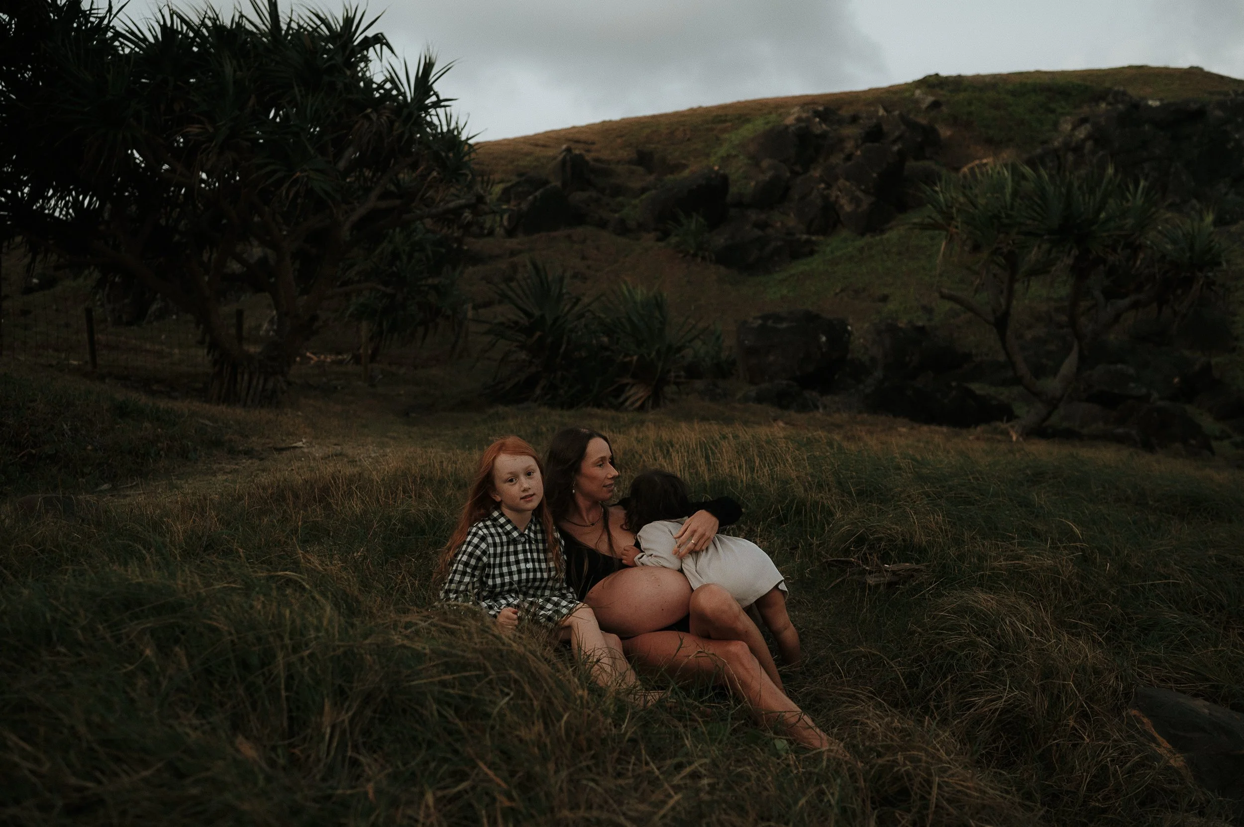 A woman with two young girls sitting on grass outdoors near large bushes and rocky hills, with a cloudy sky overhead.