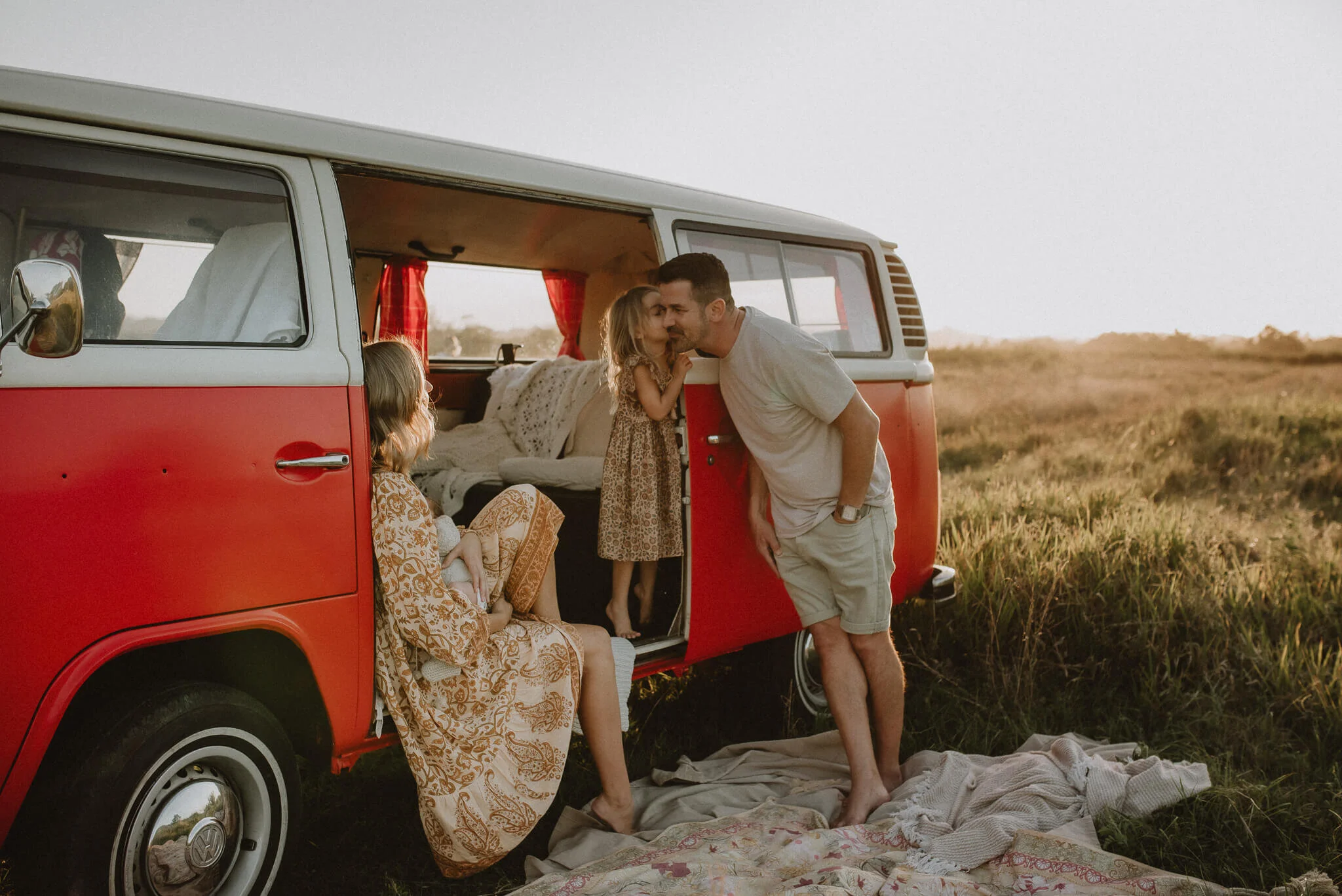 A family of four enjoying a moment outside their red and white camper van in a grassy field during sunset.