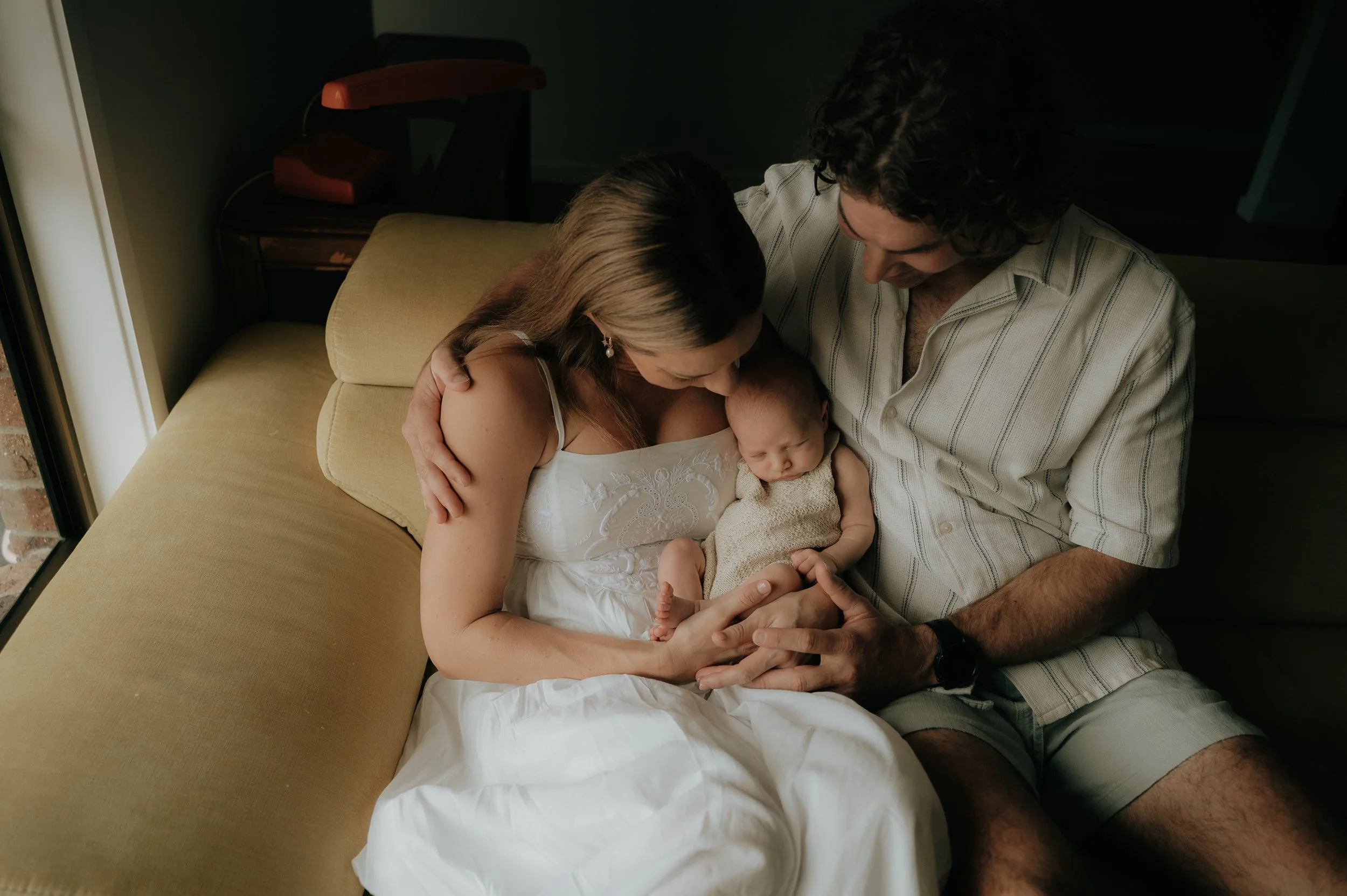 A family sitting on a beige couch, holding a newborn baby. The mother in a white dress and the father in a short-sleeve shirt and shorts are gently cradling the baby.