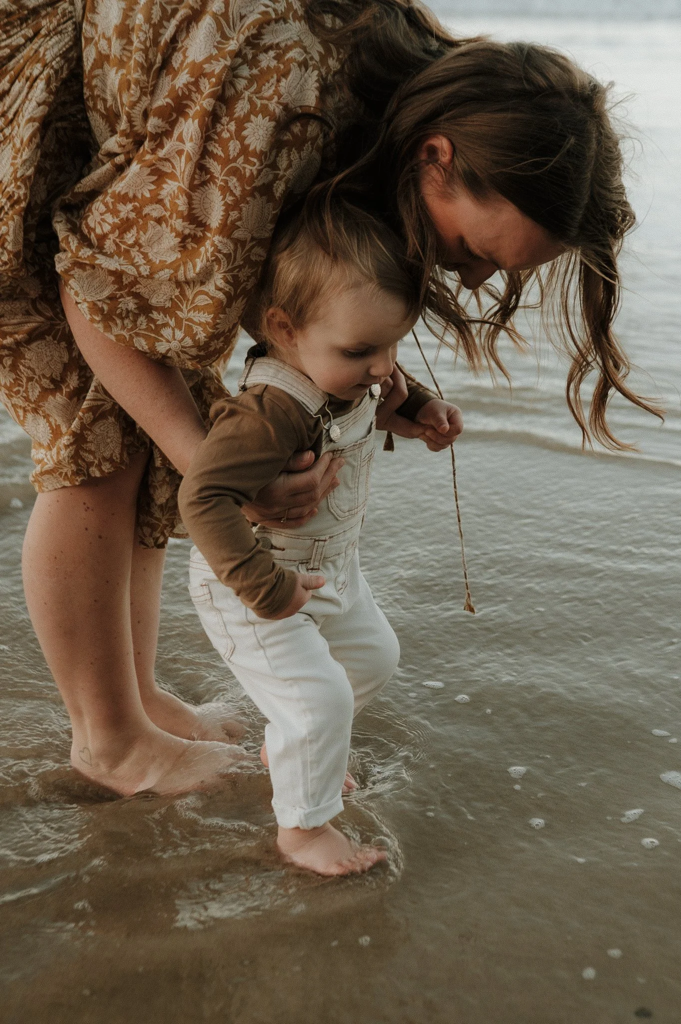 A woman and a young child wading in the ocean at the beach, with the woman helping the child stand in the water.