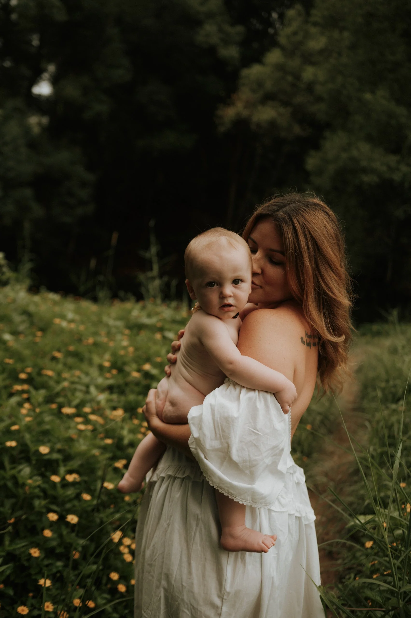 A woman holding a baby in a field of yellow flowers with trees in the background.