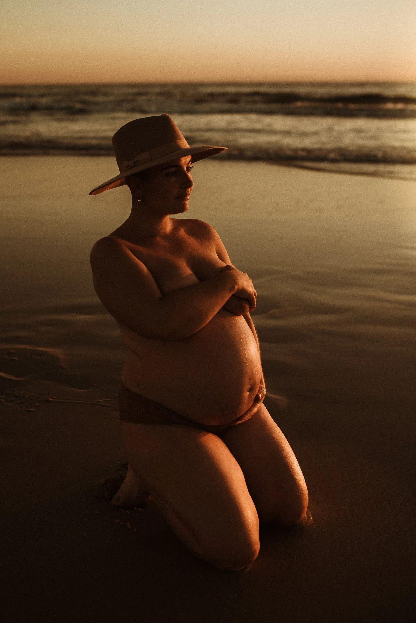 Pregnant woman wearing a hat kneeling on a beach at sunset