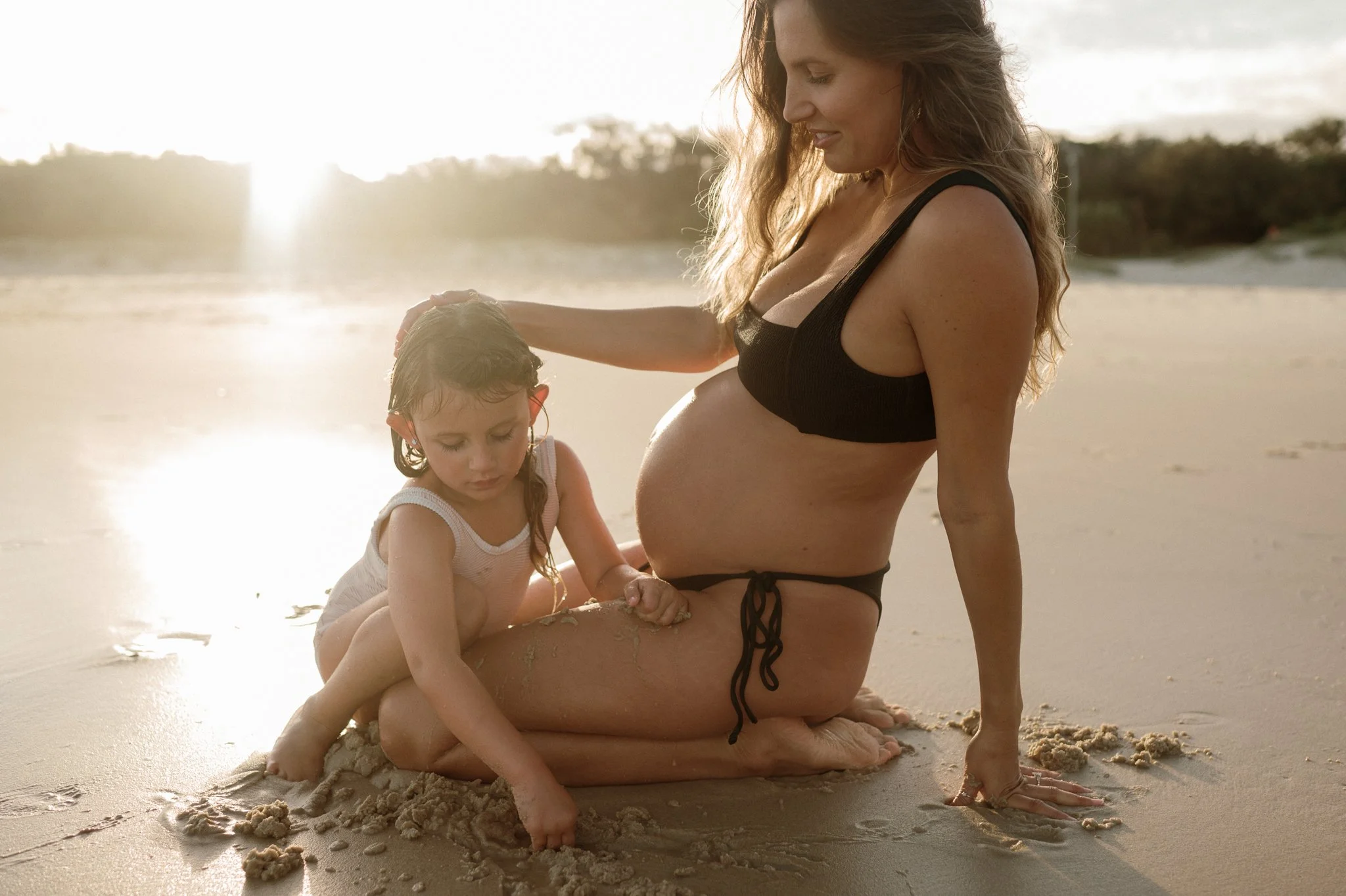 A pregnant woman in a black bikini sitting on the sand at the beach, playing with a young girl in a white swimsuit, with the sun setting in the background.