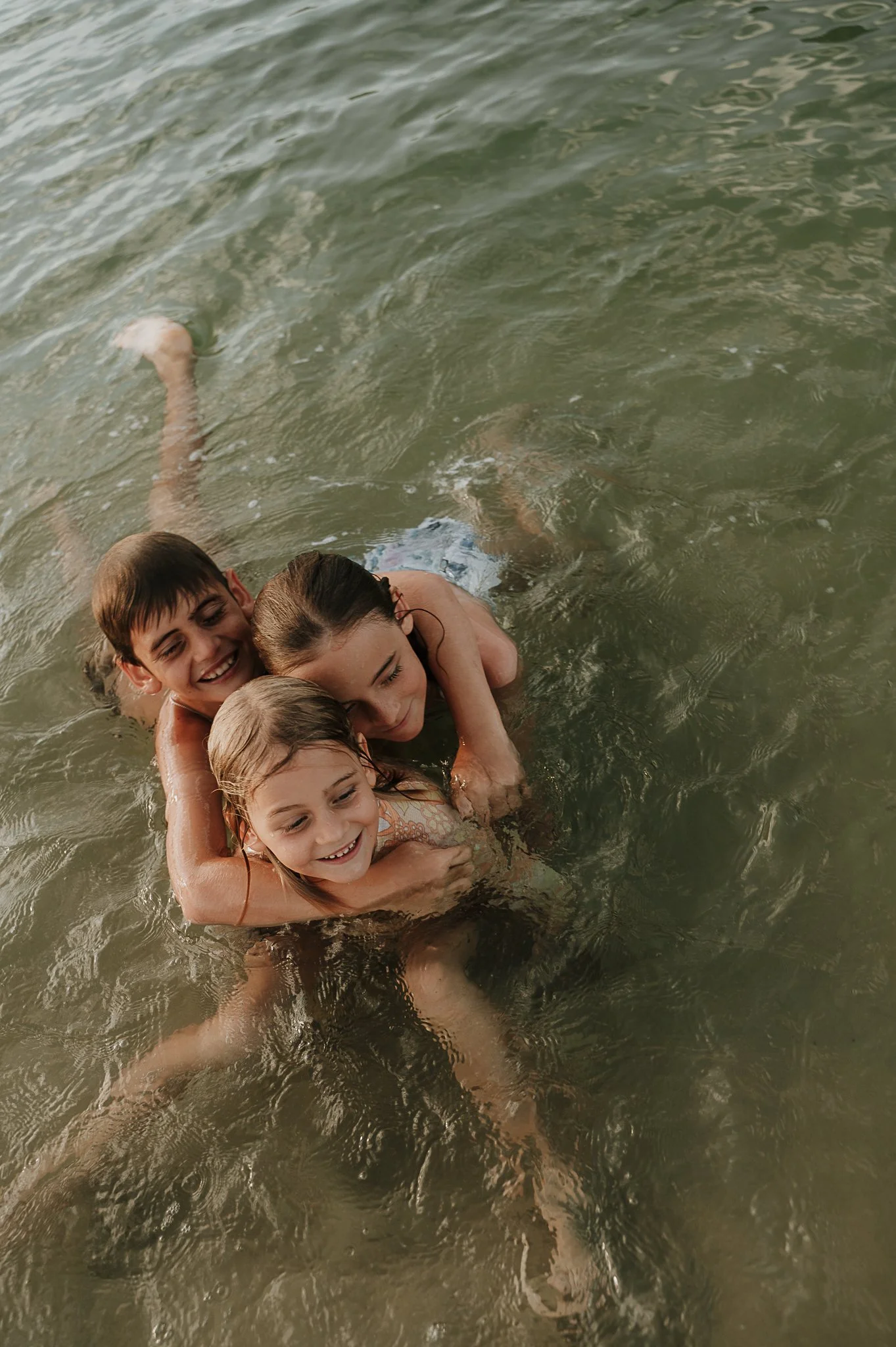 Three children are playing and hugging each other in a body of water, smiling and enjoying their time.
