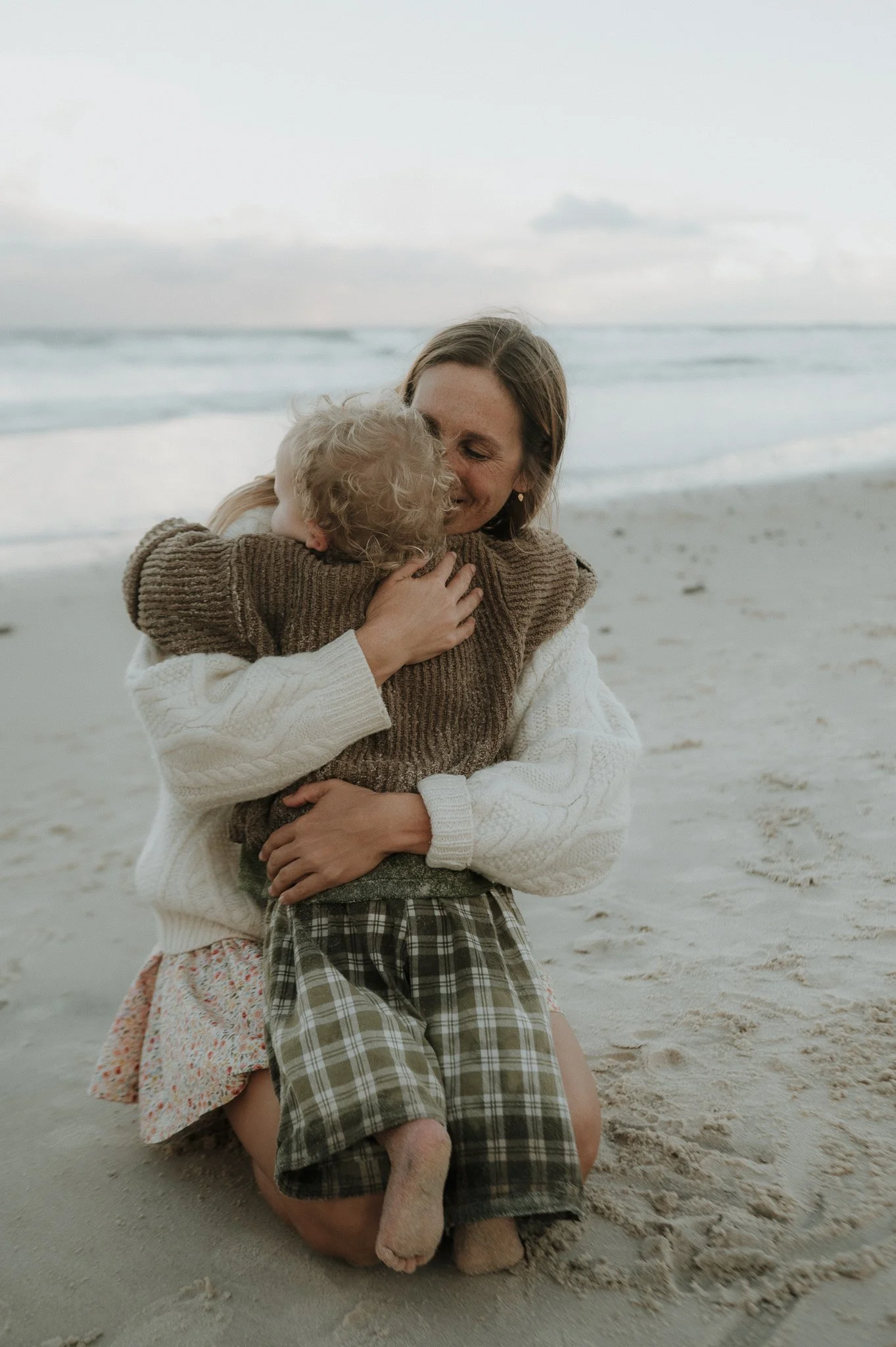 A woman and two children hugging on a sandy beach near the ocean at sunset.
