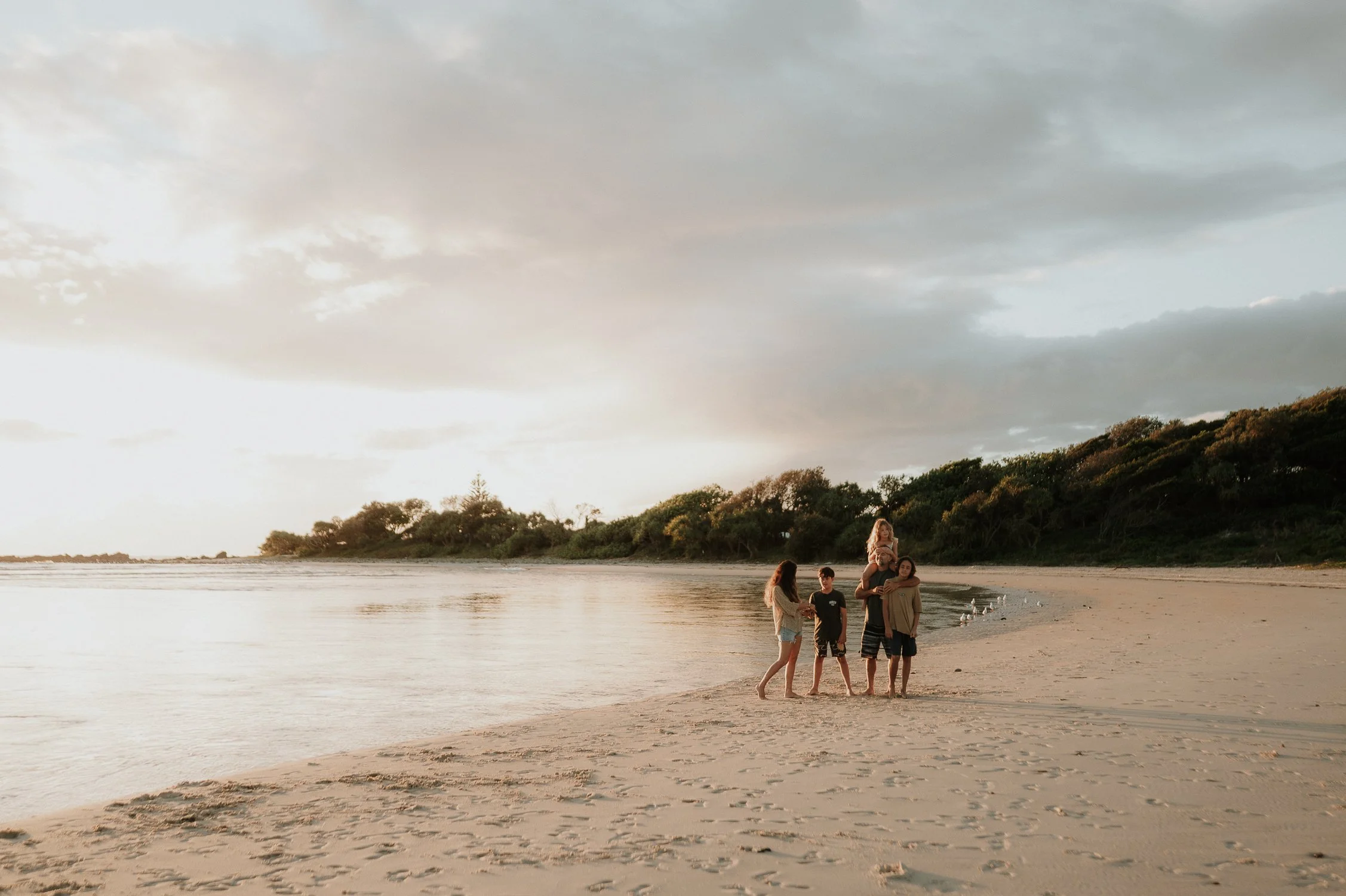A group of five people on a beach during sunset or sunrise, with two individuals on the shoulders of others, surrounded by calm water, sandy shore, and green trees in the background.