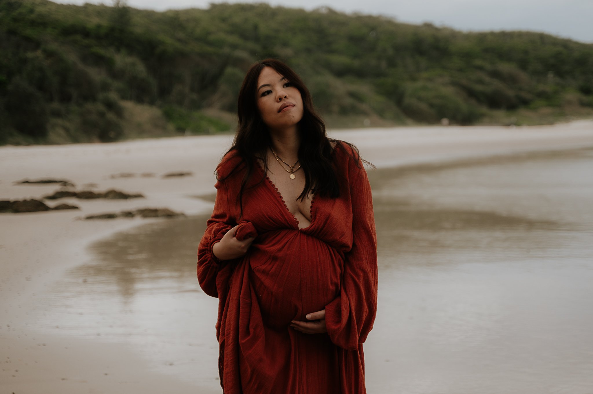 Pregnant woman in a red dress stands on the beach, holding her belly and looking thoughtful.