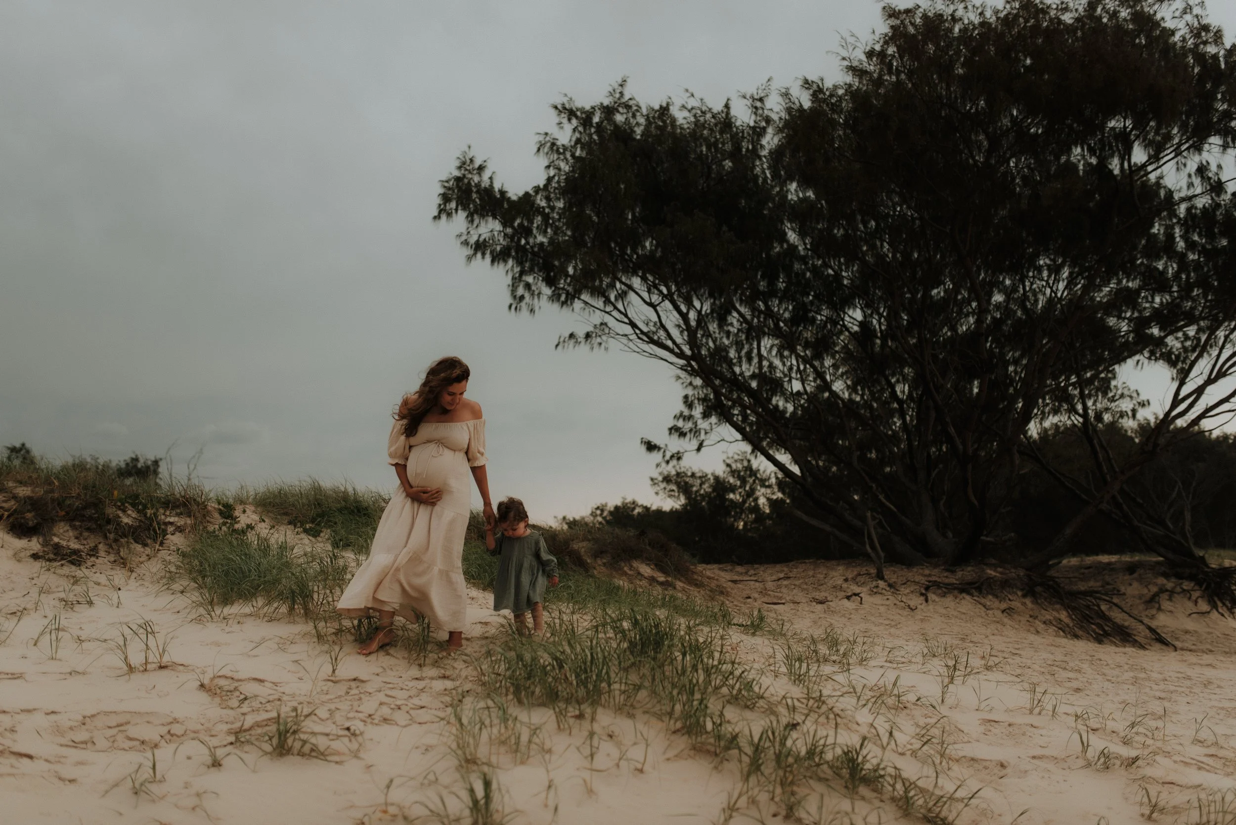 A pregnant woman in a flowing beige dress walking on the beach with a young girl, holding hands and looking down at the sand, under a cloudy sky with a large tree nearby.