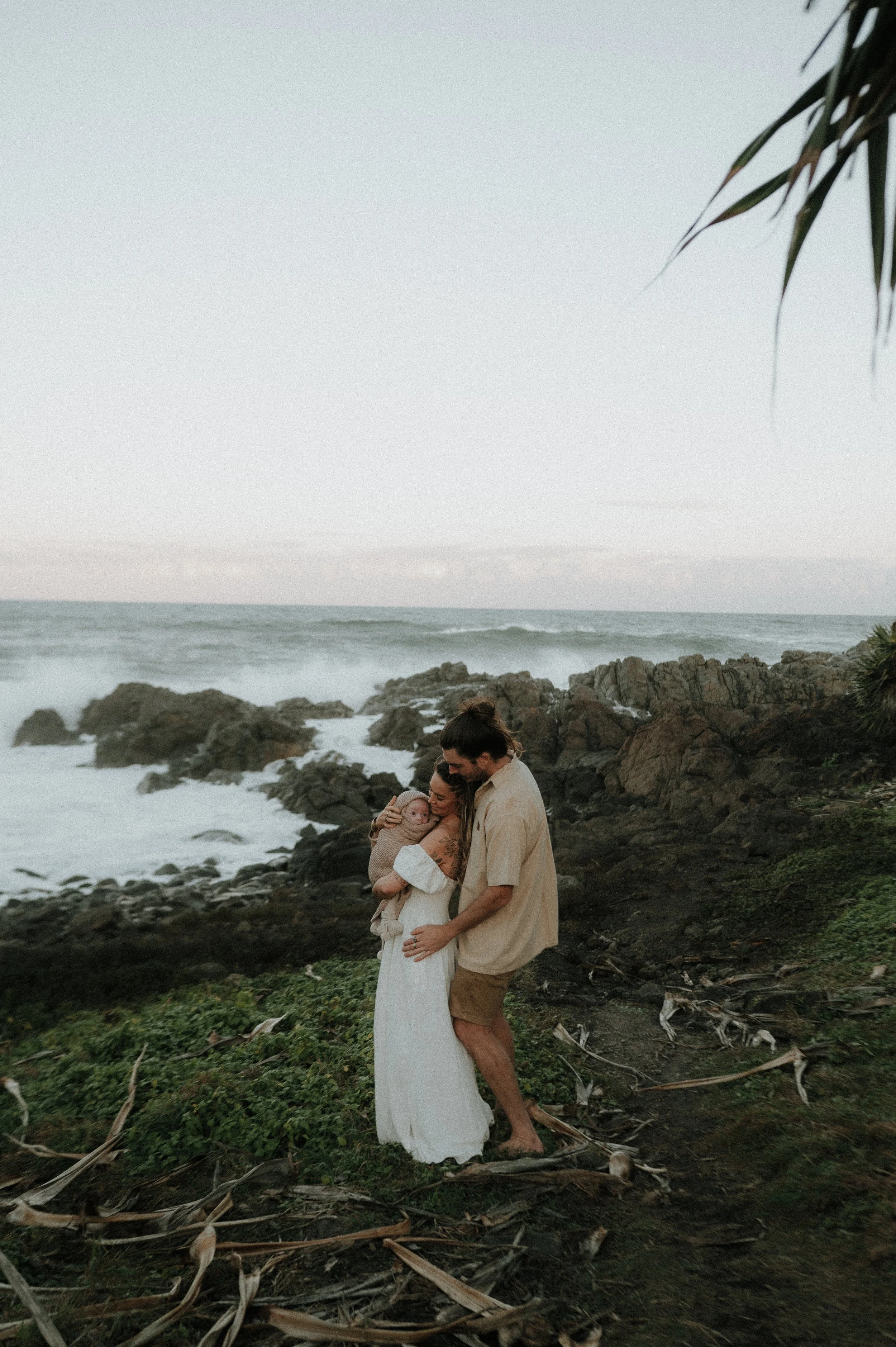 A family of three standing on a rocky beach with the ocean in the background, embracing each other during sunset.