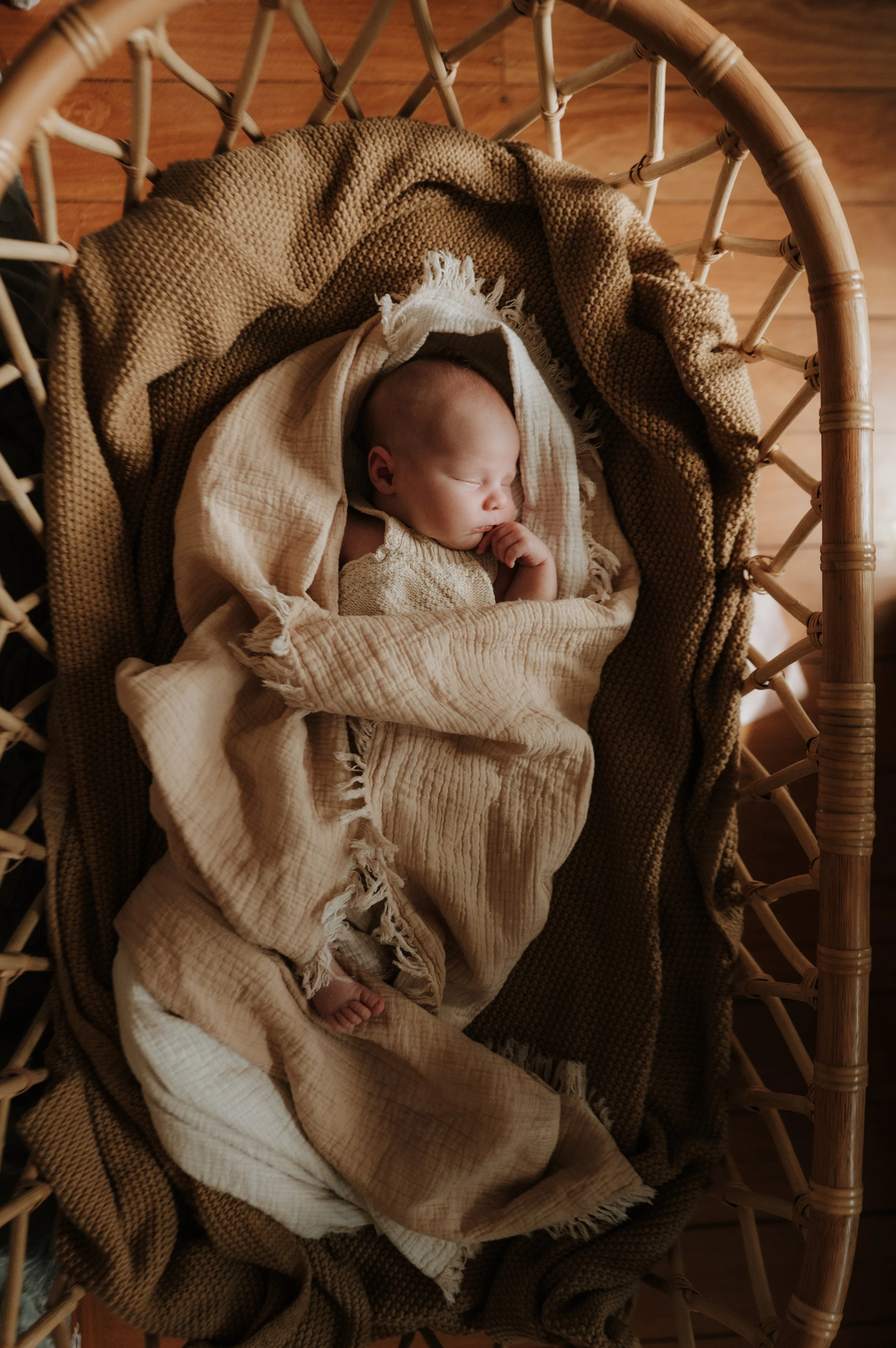 Young baby sleeping peacefully in a woven bassinet, wrapped in beige and tan blankets.