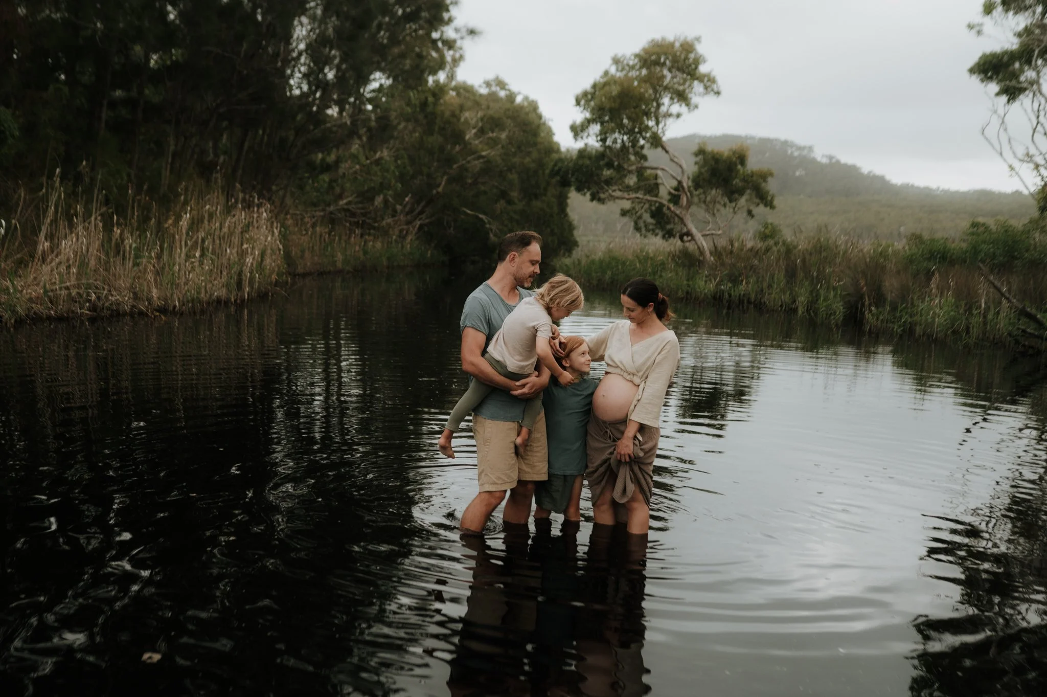 A family of four standing in a river surrounded by trees and greenery, with a pregnant woman, a man, and two children.