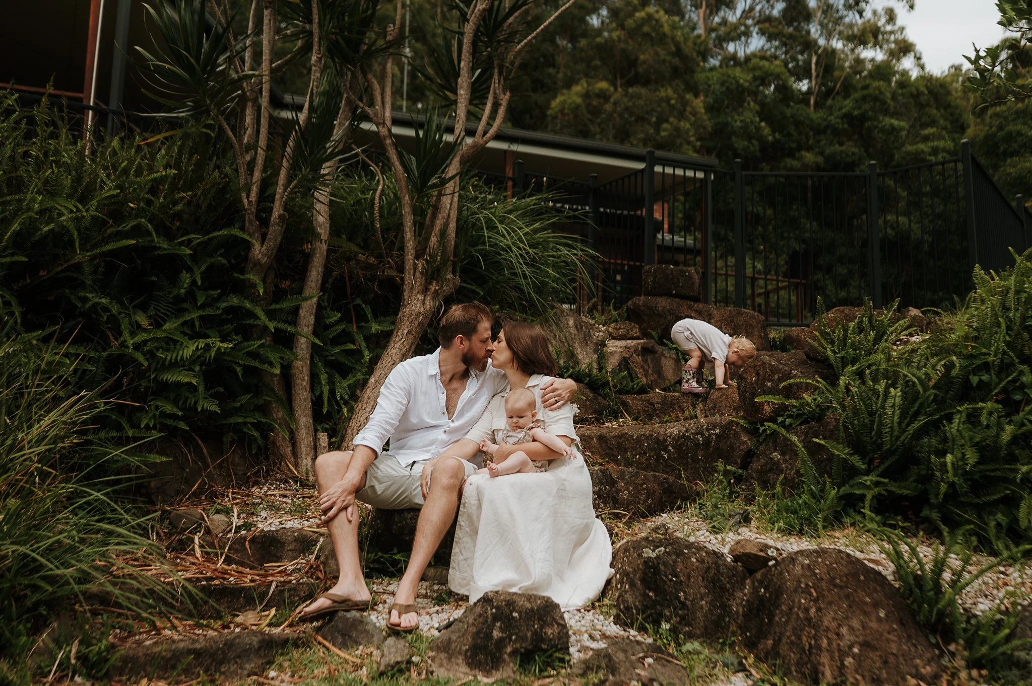 A family sitting on rocks in a natural outdoor setting with lush greenery, two adults and a baby, with a young child climbing the rocks nearby.