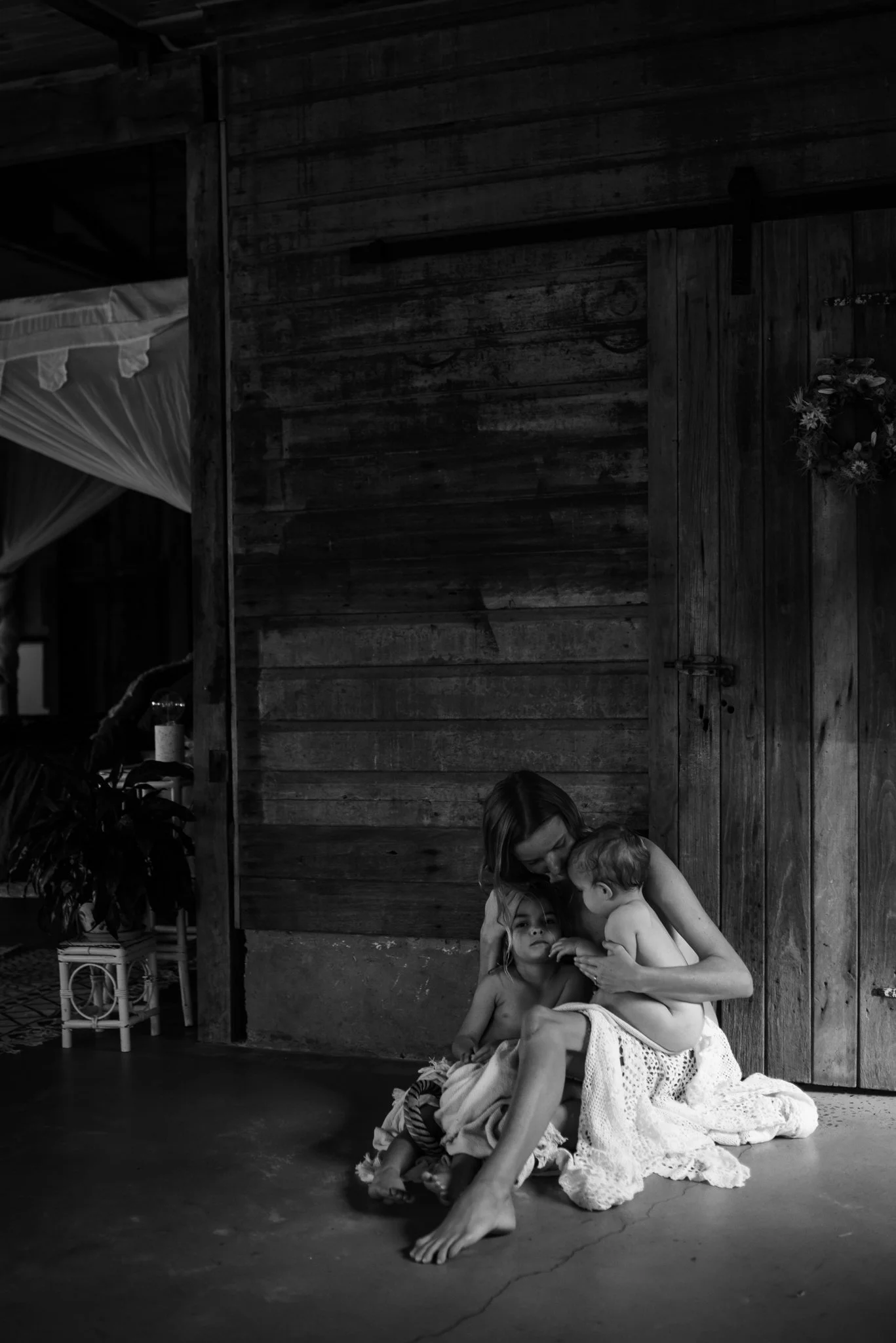 Black and white photo of a woman sitting on the floor with two young children, wrapped in a blanket, against a rustic wooden wall.