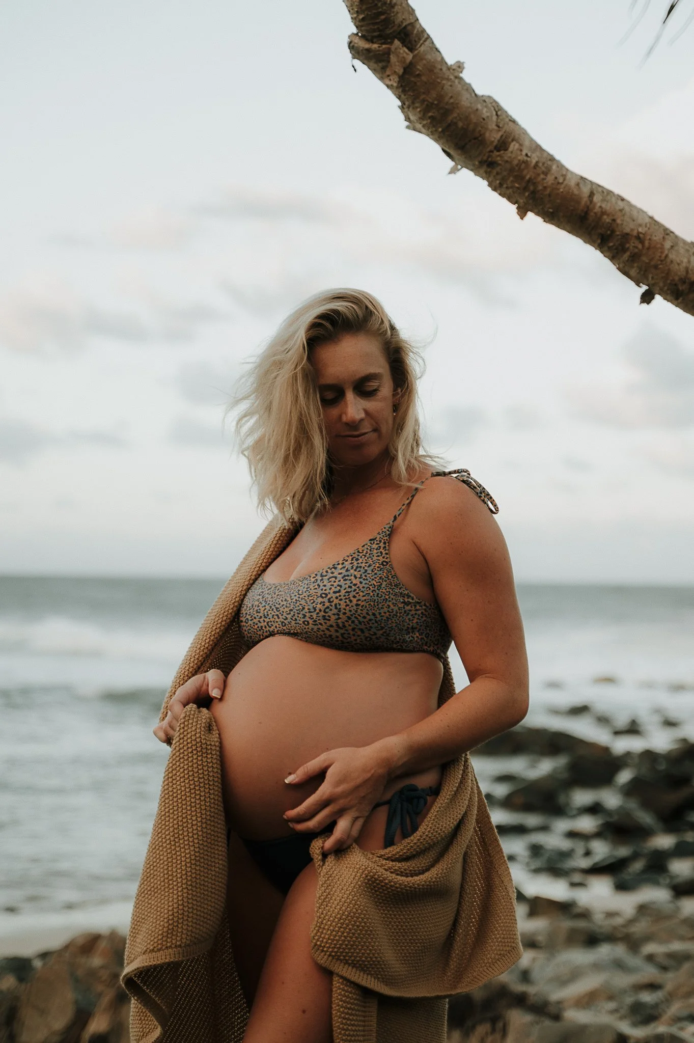 Pregnant woman in a leopard-print bikini and black shorts standing on a rocky beach, holding her belly with one hand, a brown cardigan draped over her shoulders, under a cloudy sky with a tree branch overhead.