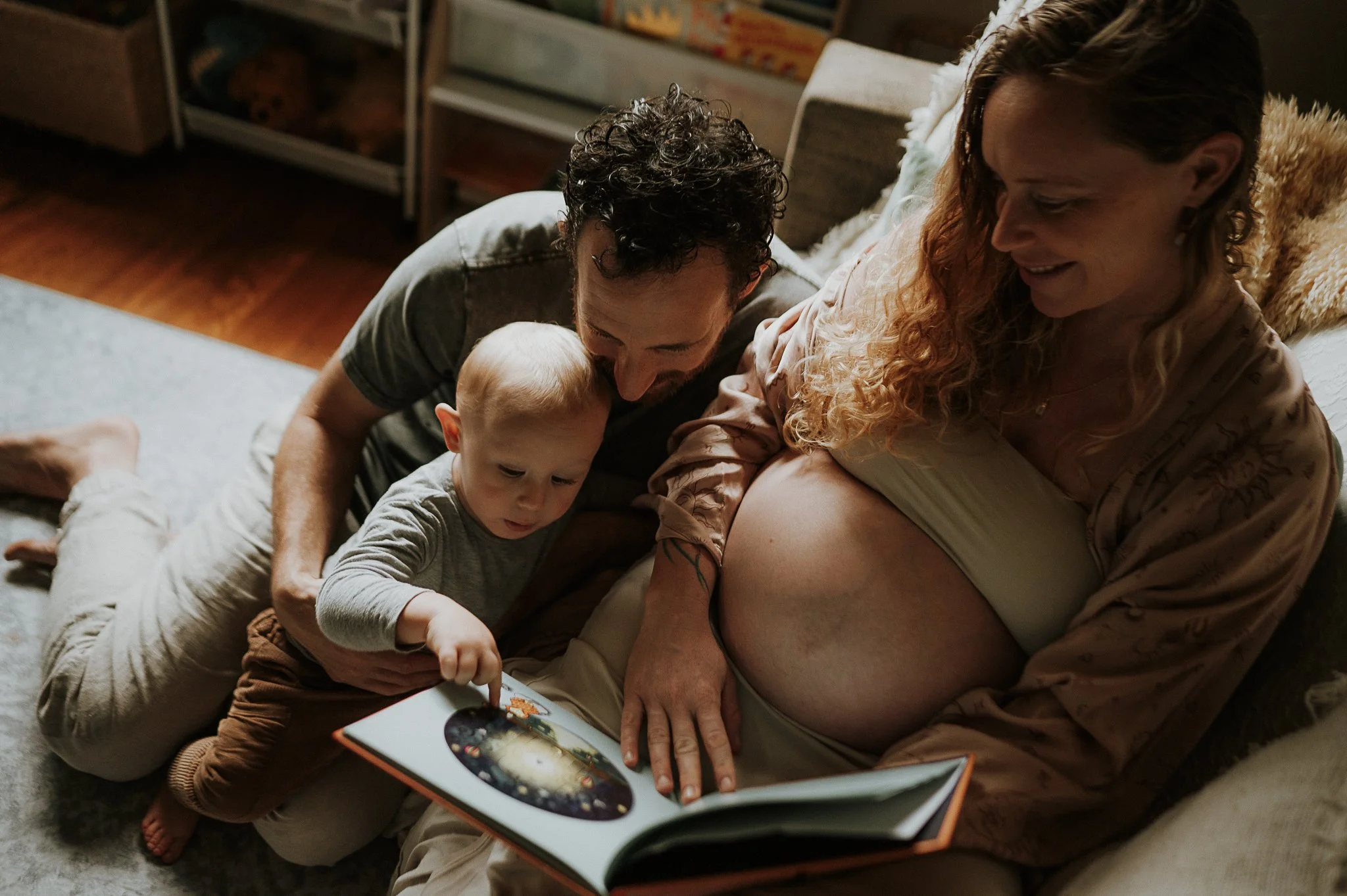 A family scene with a pregnant woman sitting on a sofa, a man and a young boy reading a book together, in a cozy living room.