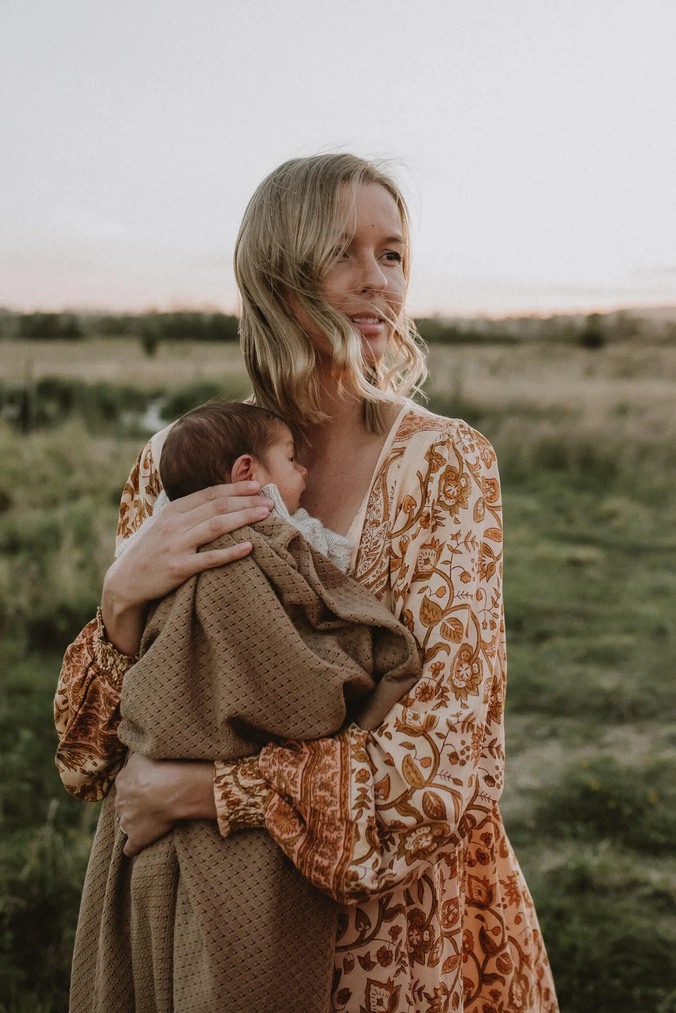 A woman with blonde wavy hair holding a sleeping baby wrapped in a brown blanket in an outdoor field at sunset.