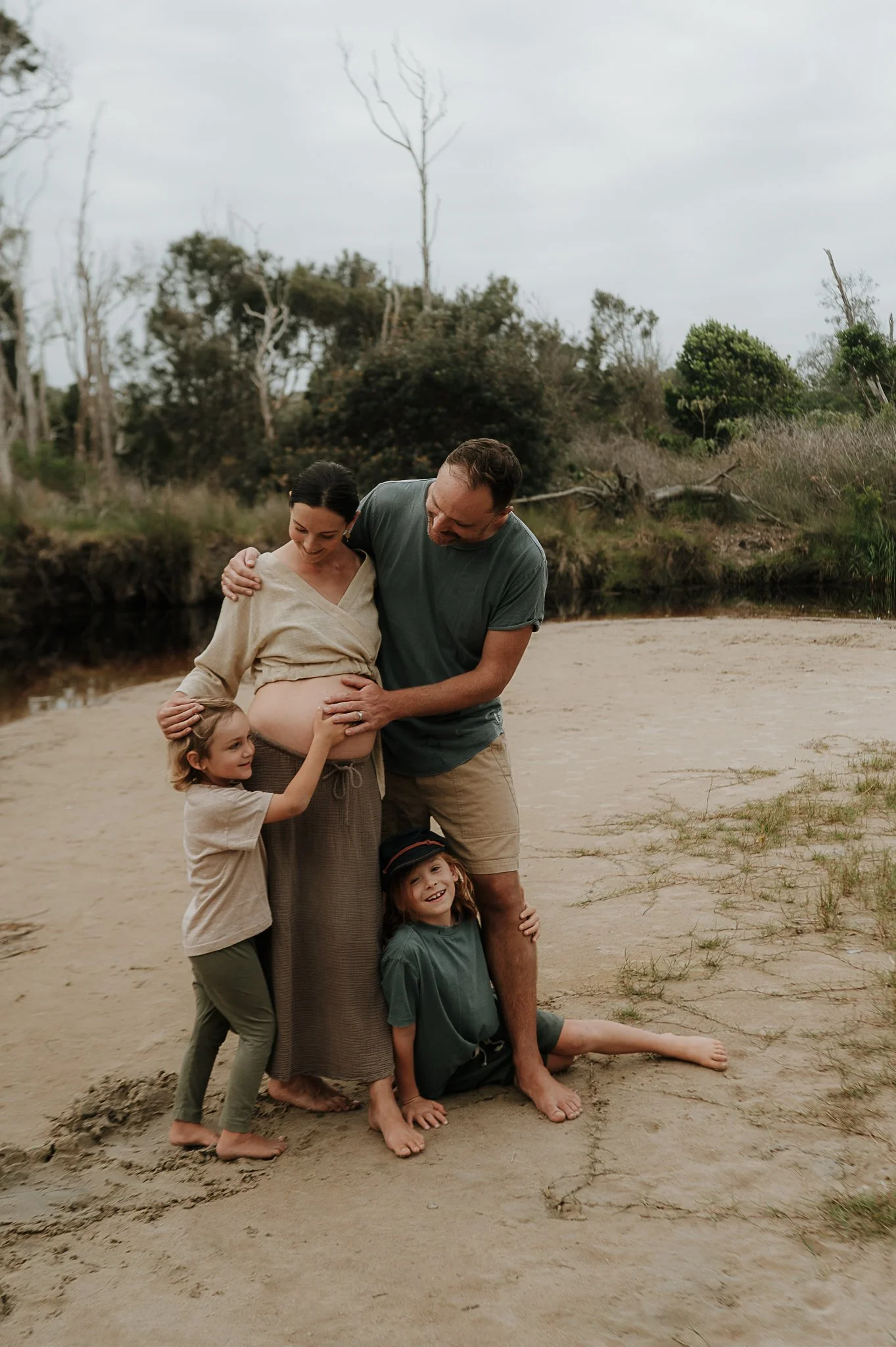 A family of five, including a pregnant woman, a man, and three children, gathered on a sandy beach during overcast weather, with trees and shrubs in the background.