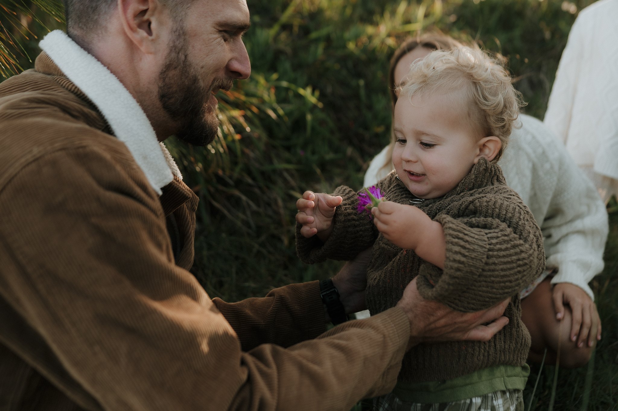 A father and young child sharing a moment outdoors, with the child holding a purple flower.