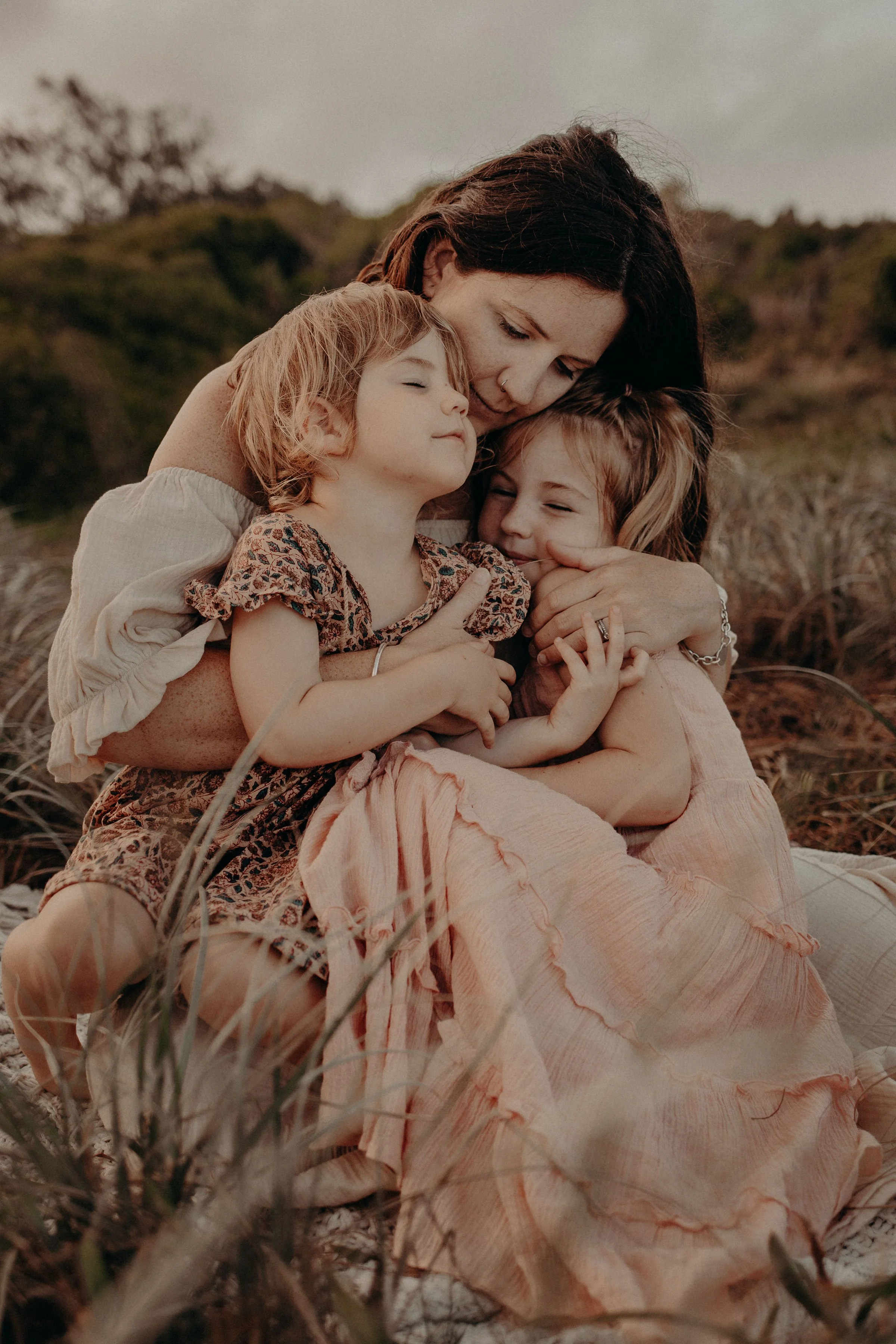 A woman embraces two young girls outdoors in a natural setting with grass and trees, during the daytime.