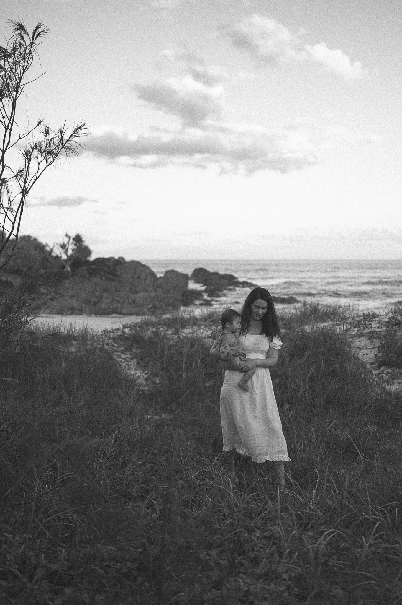 Black and white photo of a woman holding a baby by the seaside, surrounded by grass and rocks, with an ocean backdrop and cloudy sky.