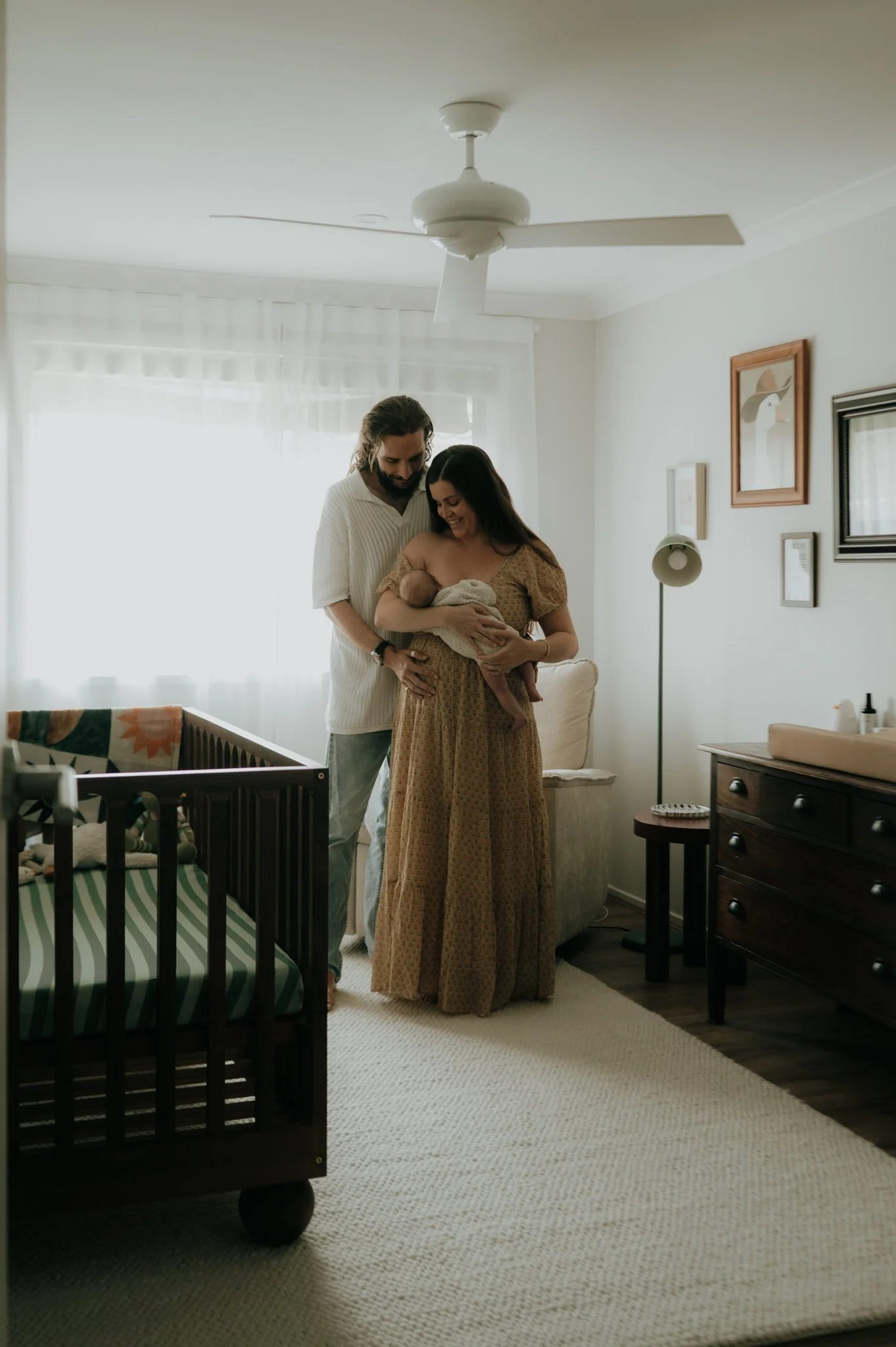 A family in a bedroom, with a woman holding a newborn baby and a man standing beside her, both smiling and looking at the baby.