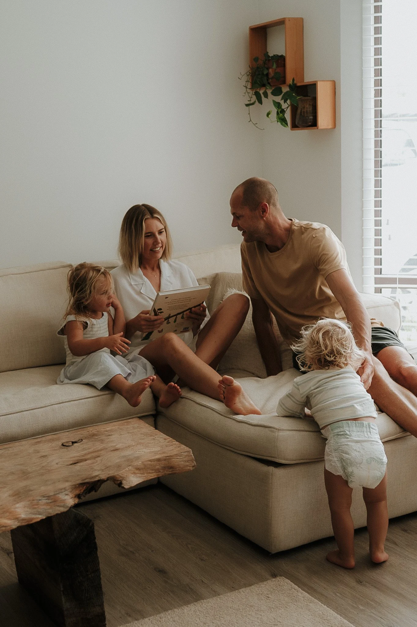 A family sitting on a beige sofa in a bright living room, with two young children and a woman reading a book, while a man and a toddler play on the floor nearby.