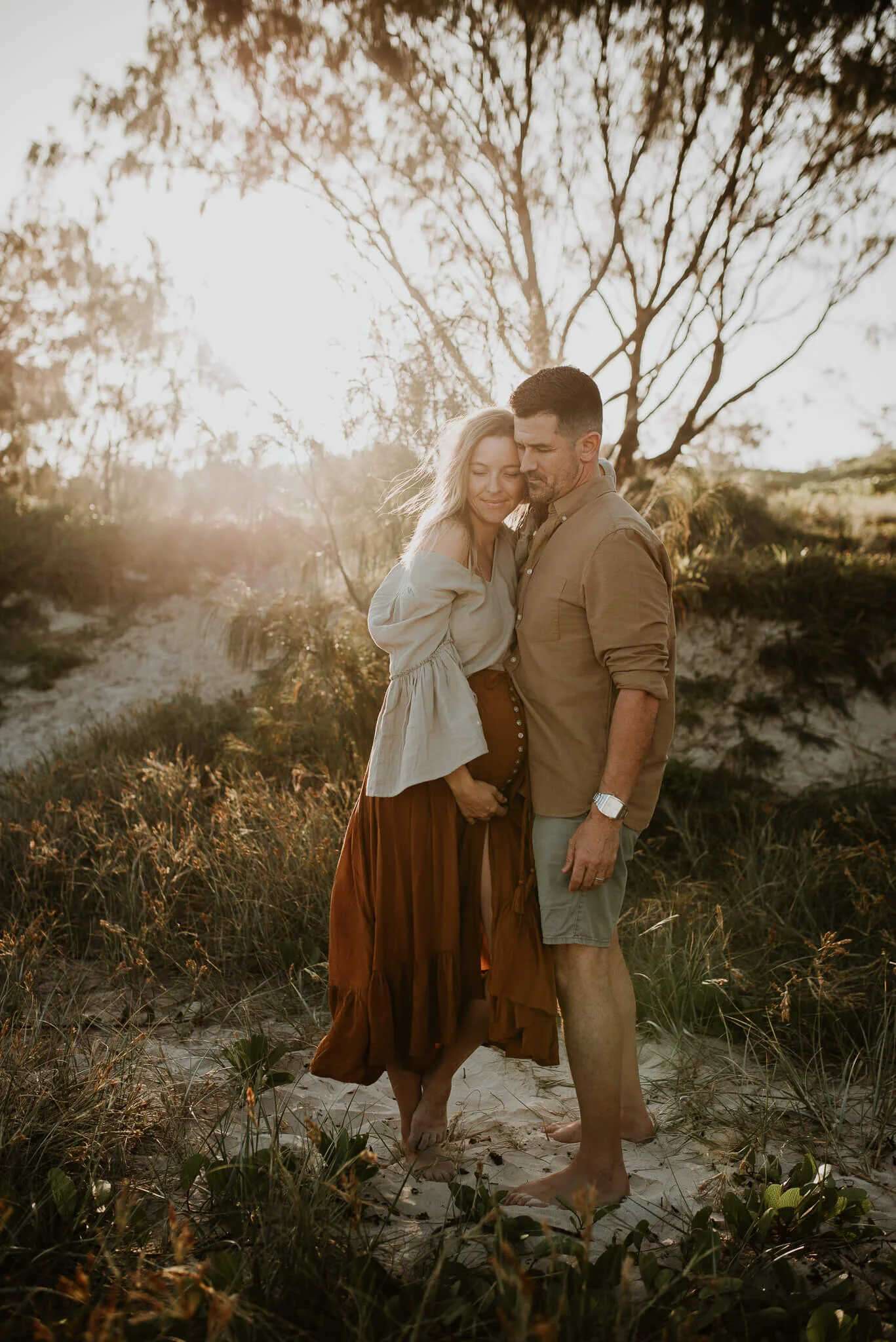 A couple standing close together outdoors at sunset, surrounded by grassy dunes and trees, with warm light illuminating their faces.