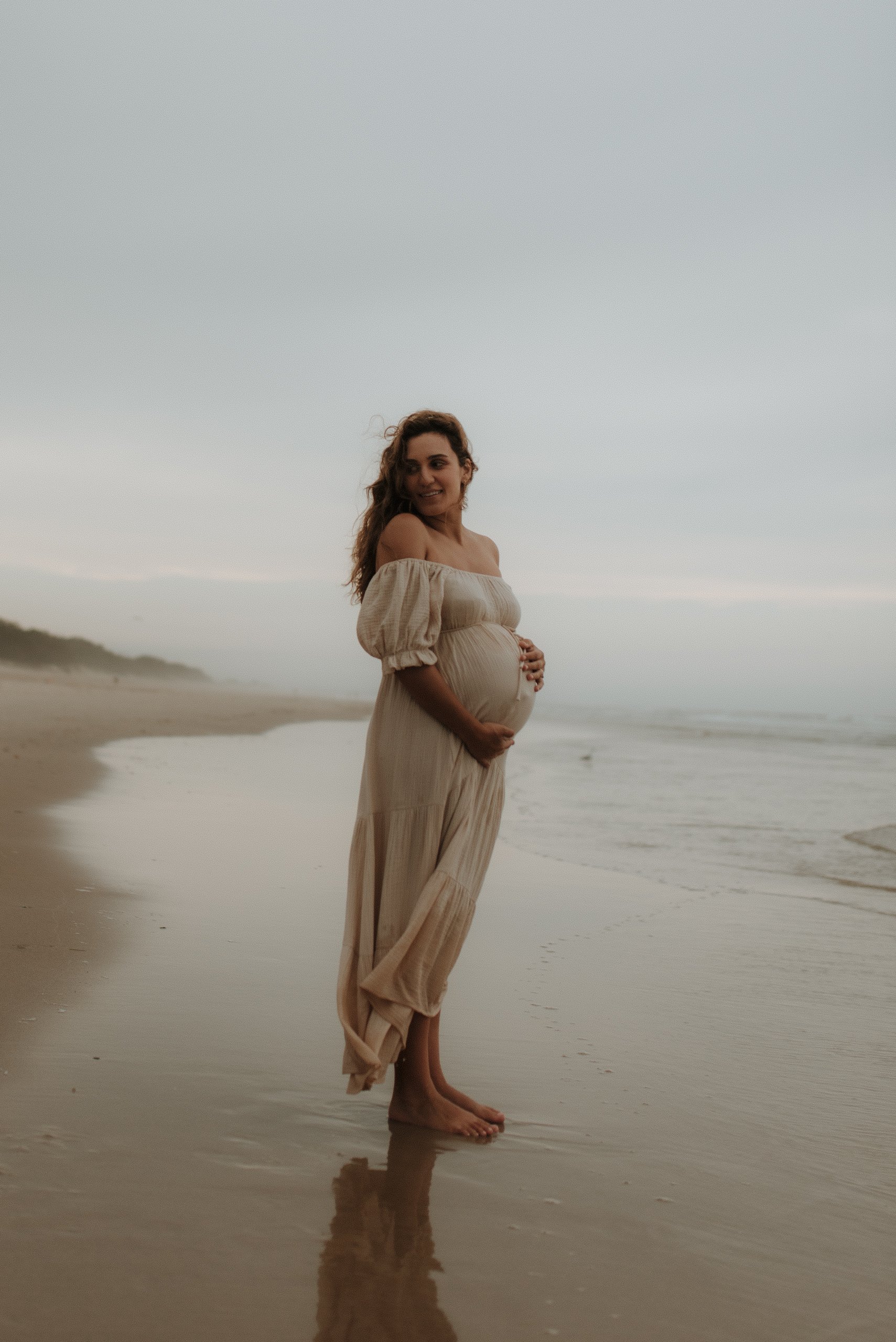 Pregnant woman in a flowing beige dress standing barefoot on the beach, holding her belly, with the ocean and cloudy sky in the background.