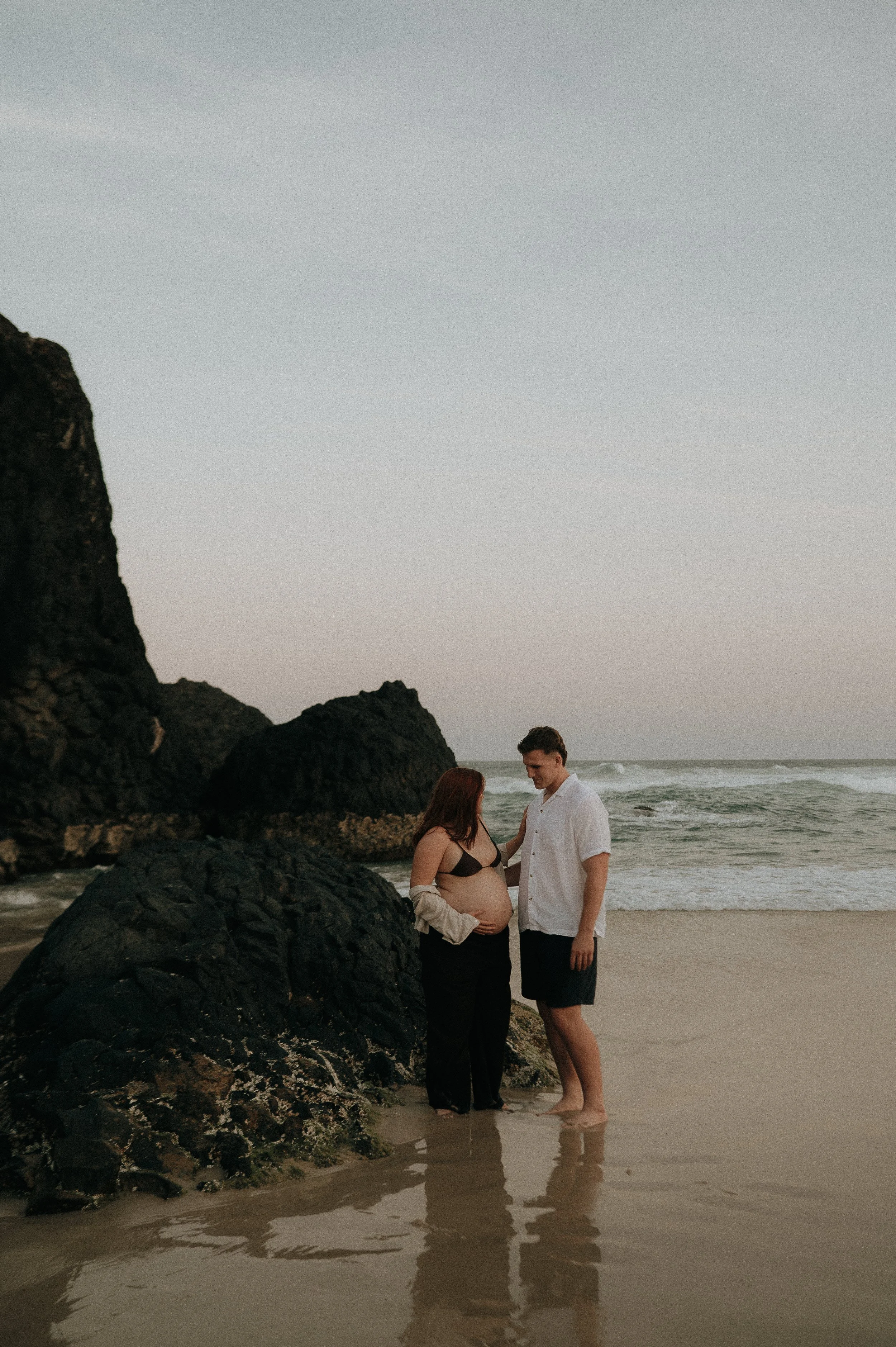 A pregnant woman and a man standing on a beach near large rocks, with the ocean and sky in the background, during sunset.