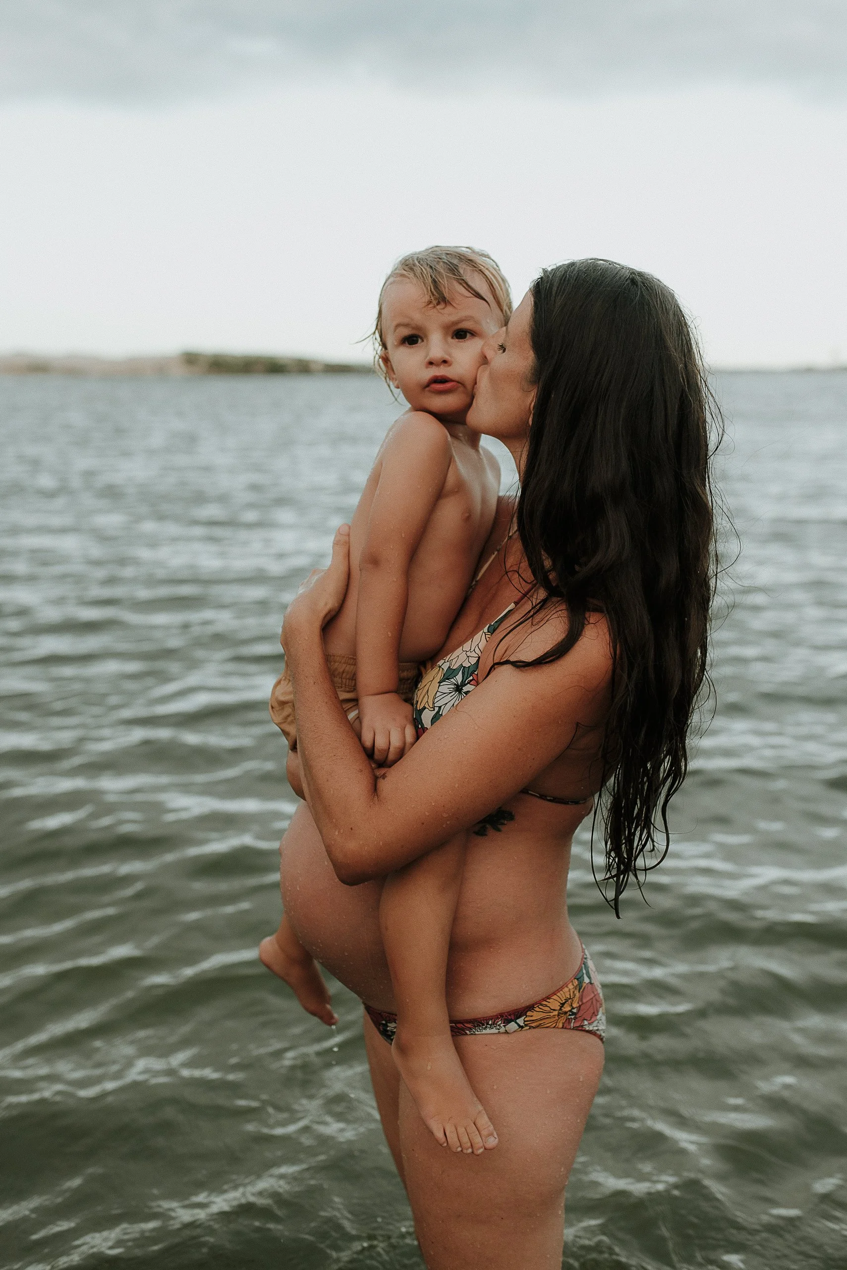 A woman with long dark hair, in a floral swimsuit, holding a young boy with wet hair, giving him a kiss on the cheek while standing in a body of water.