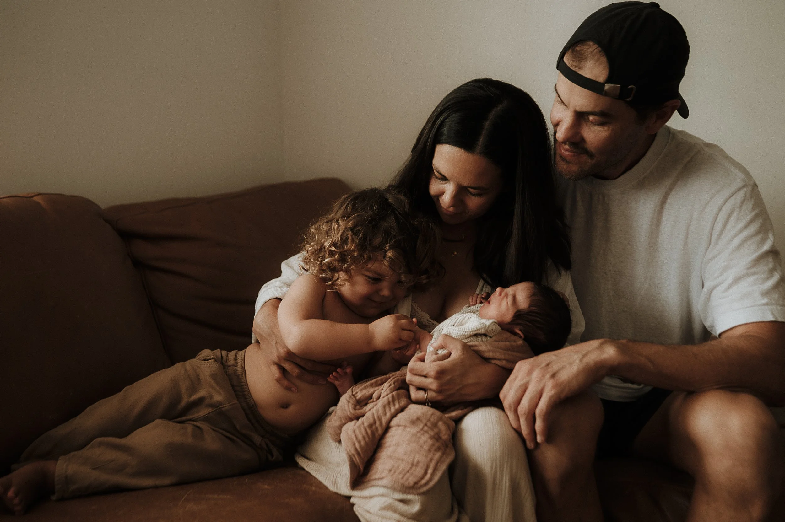 Family of four sitting on a couch, with a woman holding a newborn baby, a young boy with curly hair leaning on her, and a man wearing a cap smiling at the baby.