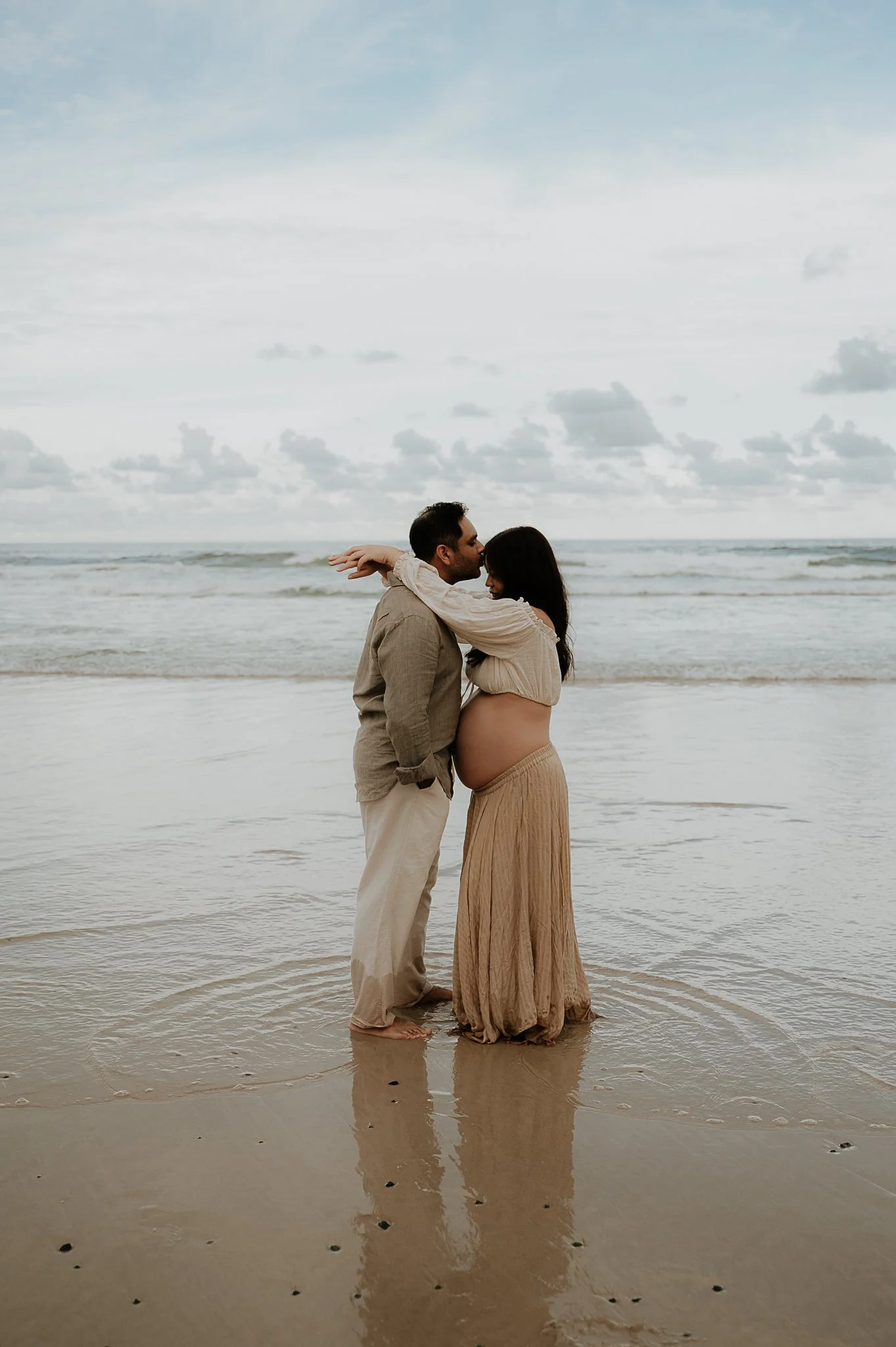 A couple standing close on the beach, with the man kissing the pregnant woman on her forehead, their feet in the wet sand near the water, under a cloudy sky.