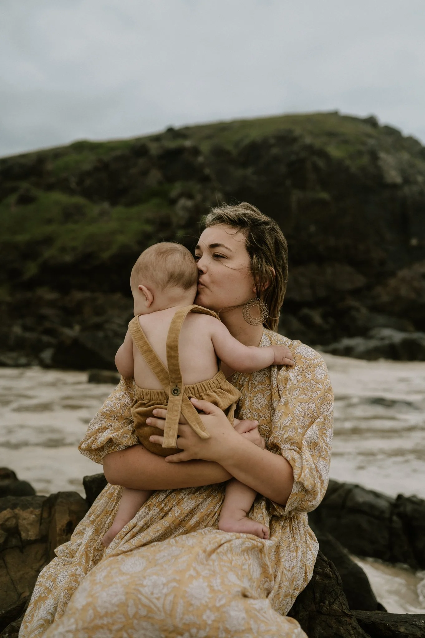 Woman holding and kissing a baby on a rocky beach with green hills in the background.