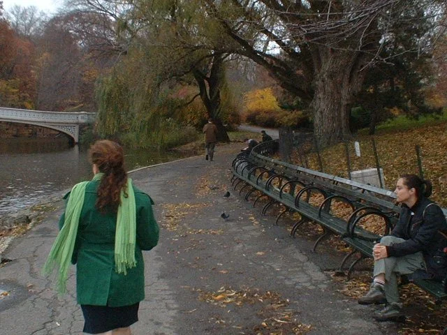 A park pathway with two women, one walking away in a green coat and bright green scarf, the other sitting on a bench, and a person walking in the distance under autumn trees.