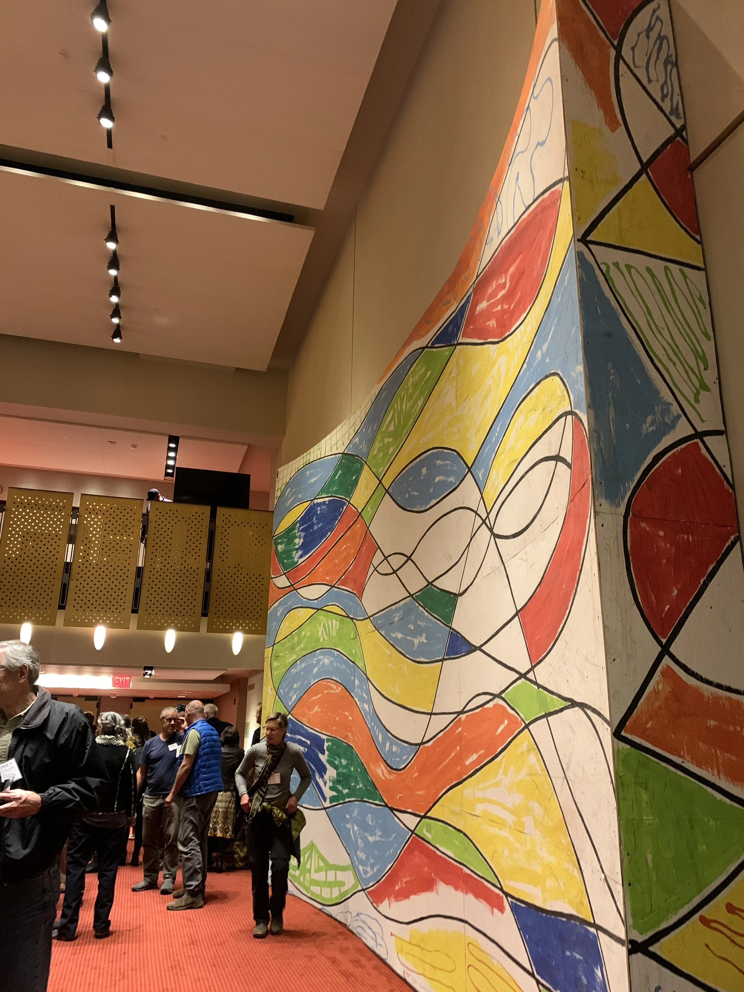 Large abstract colorful painting with various shapes and lines displayed at an indoor event, with people gathered around in the background.