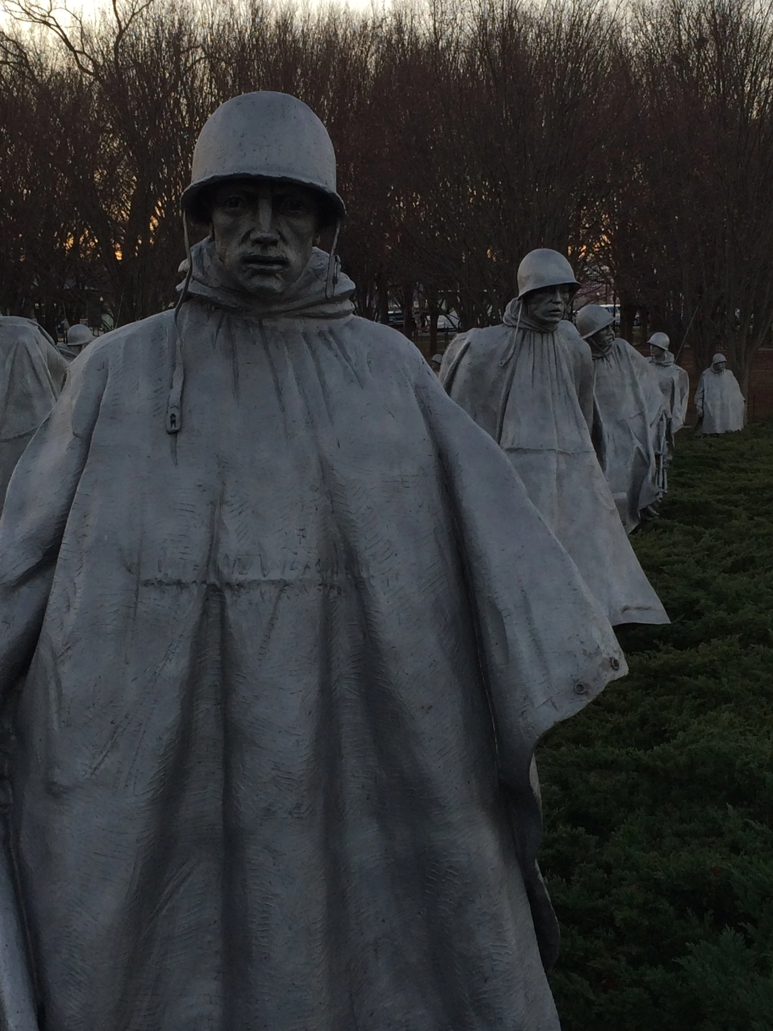 Statues of soldiers wearing helmets and cloaks lined up in a memorial or park, with a backdrop of trees at sunset.