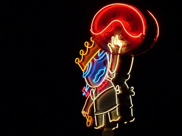 Neon sign of a girl with a large lollipop covering her face, wearing a crown and a dress against a dark background.