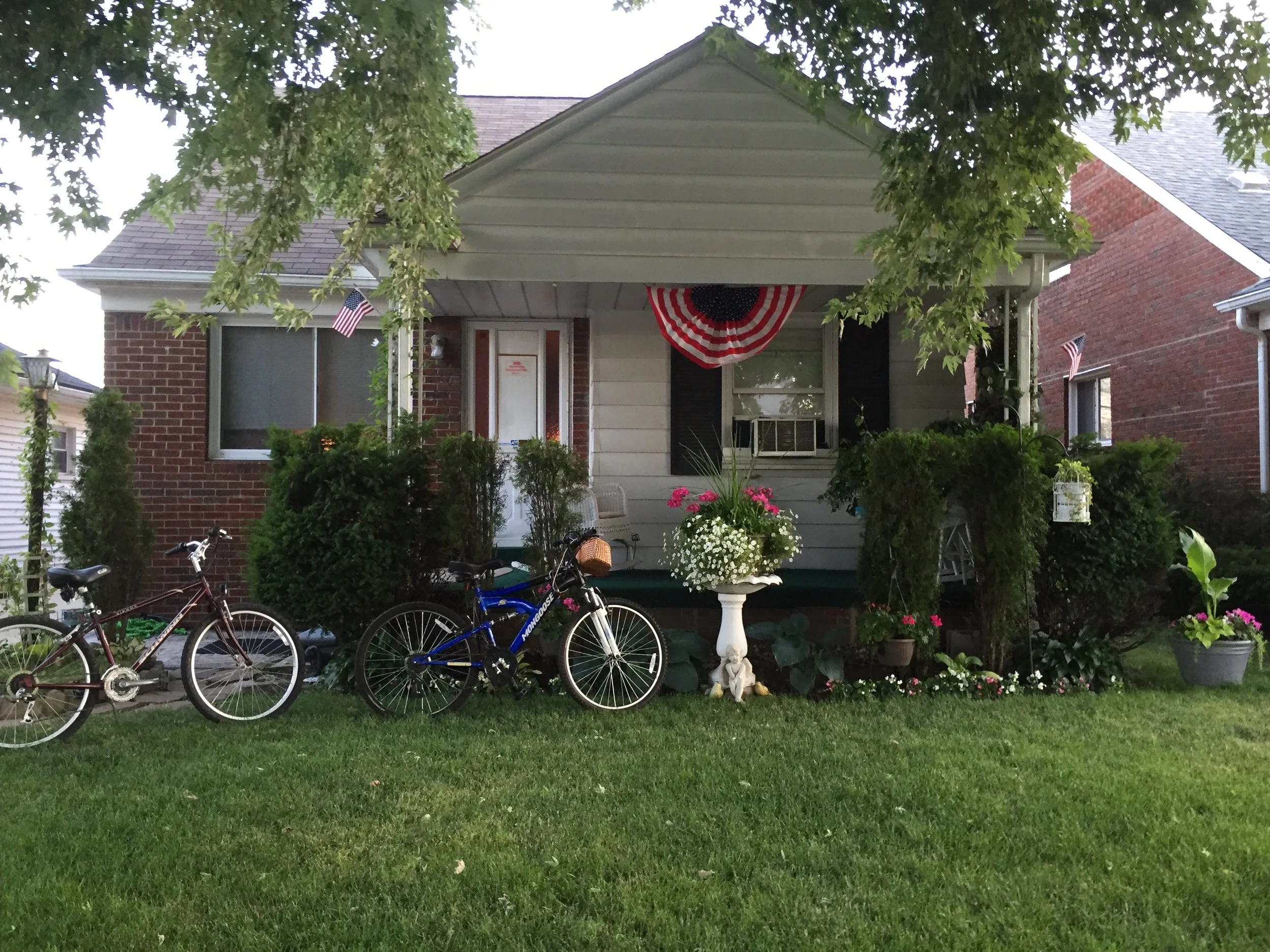 A suburban house with front porch decorated with American flags and red, white, and blue bunting, surrounded by potted plants, bicycles, and a lush green lawn.