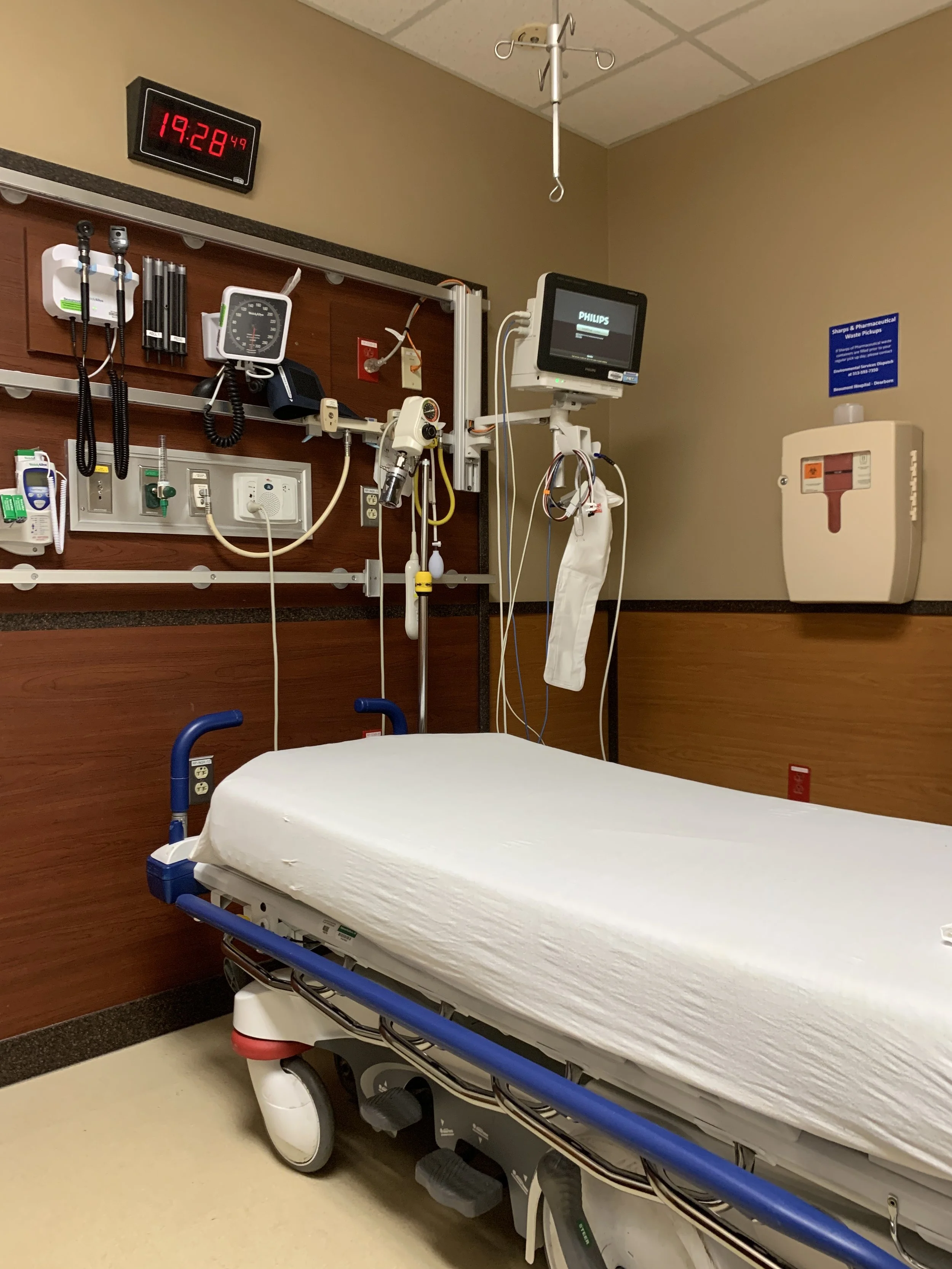 Empty hospital room with a hospital bed, medical equipment on the wall, a monitor, and a digital clock showing 19:28.