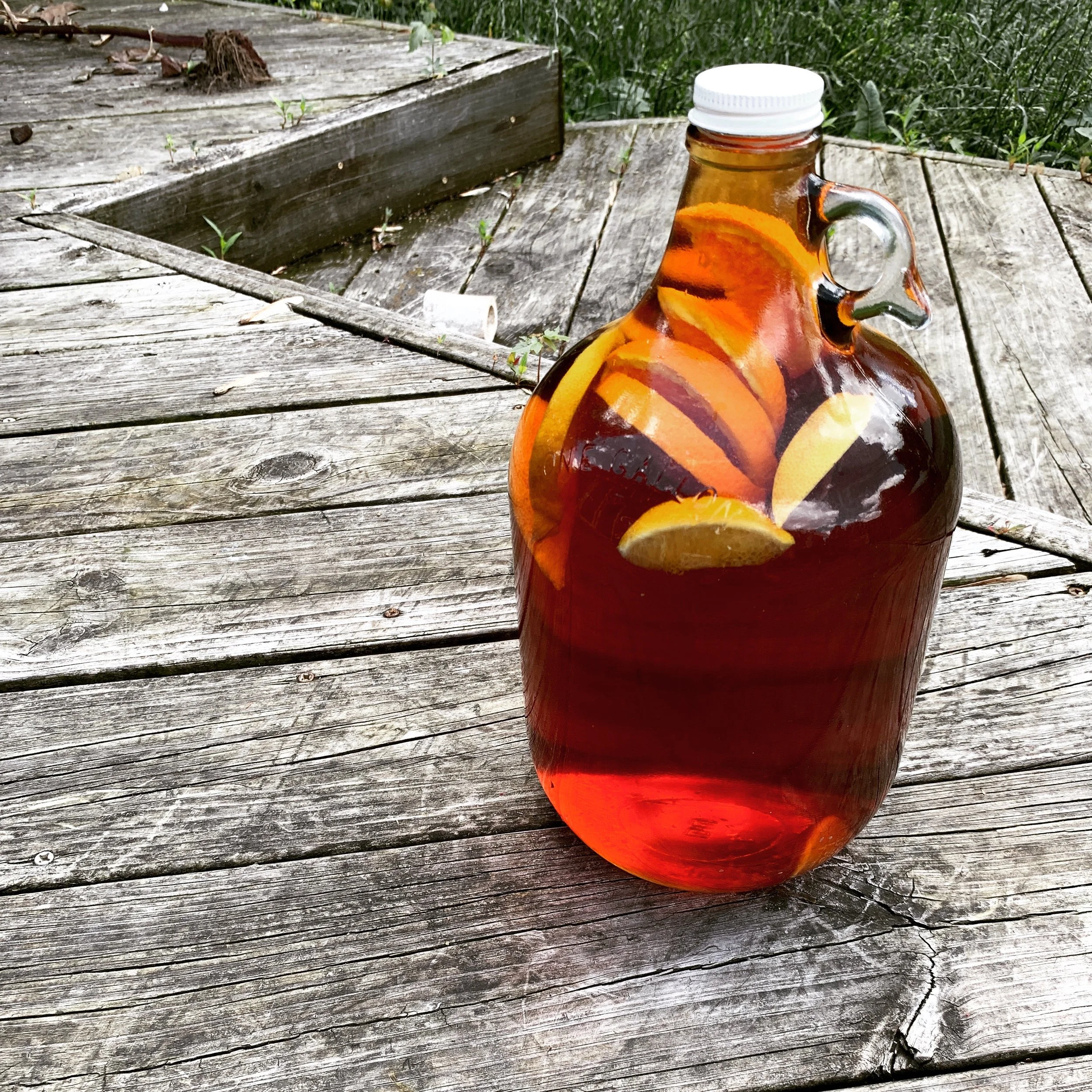 Glass jug with sliced citrus fruits, such as oranges and lemons, filled with amber-colored liquid, placed on a weathered wooden surface outdoors.