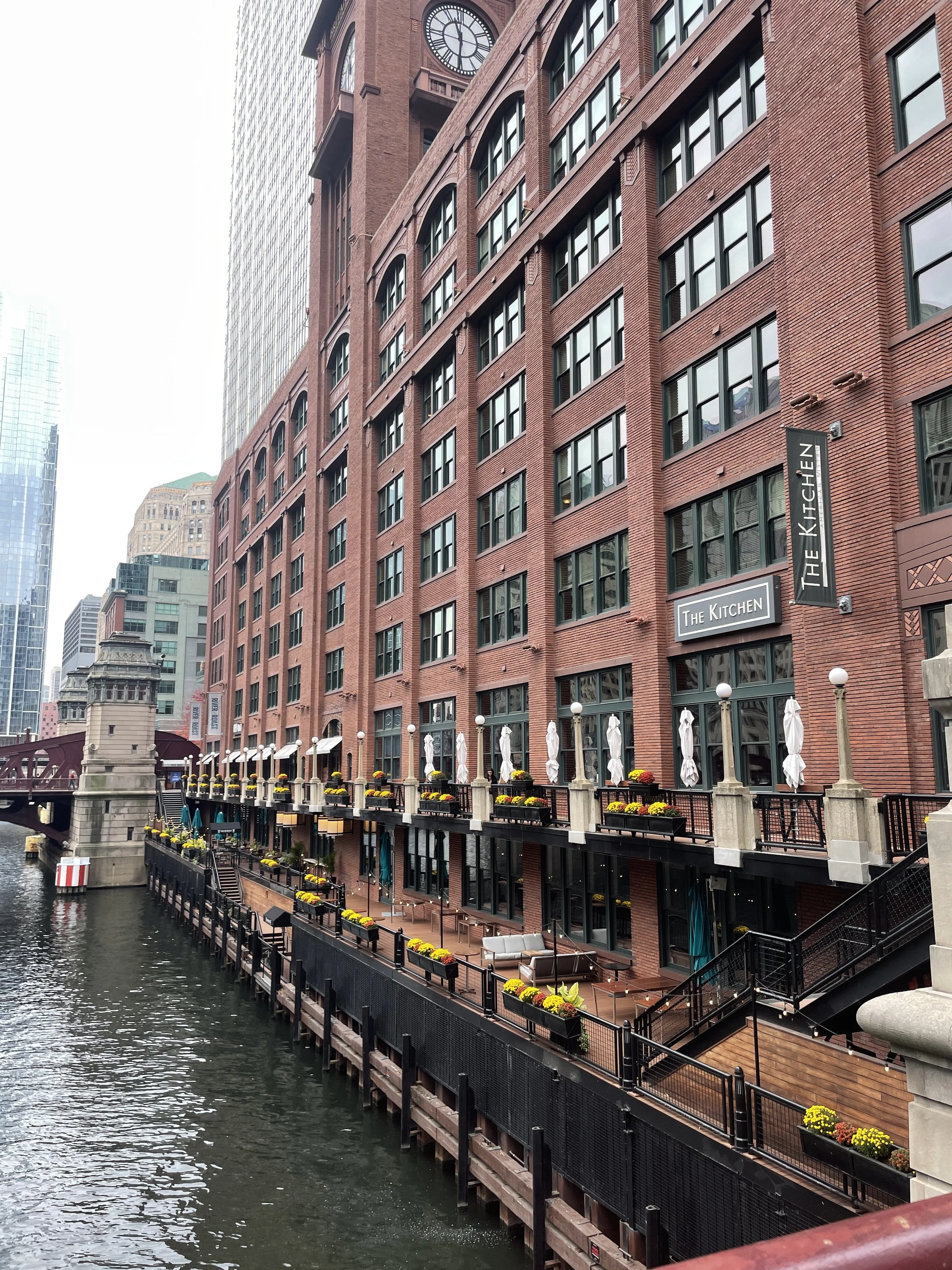 Red brick building with large windows next to a waterway, with outdoor seating area on a higher level decorated with yellow and red flowers, umbrellas, and patio furniture, and a bridge in the background.