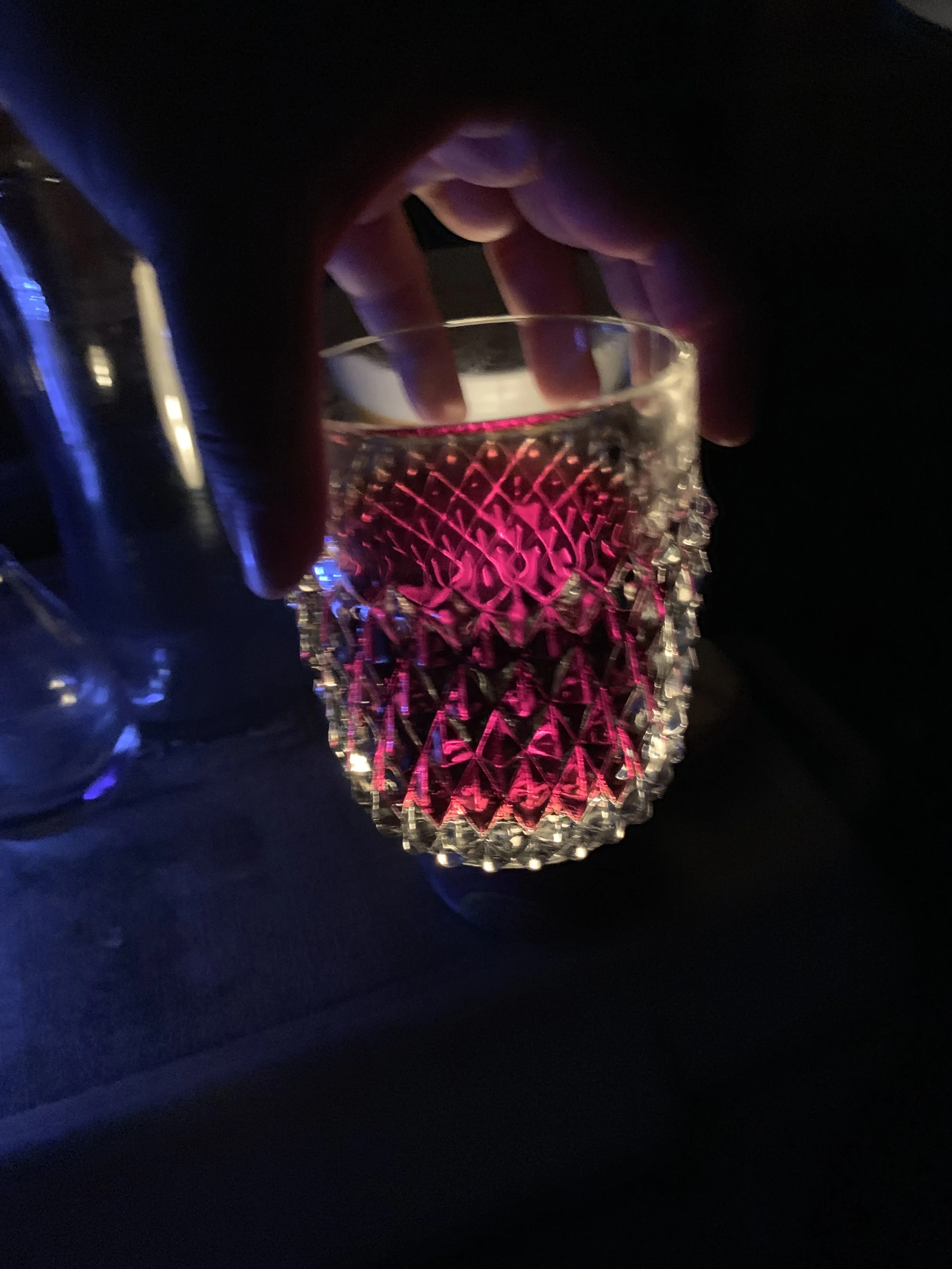 Close-up of a hand holding a glass with pink colored liquid, in a dimly lit environment with blue and pink lighting.