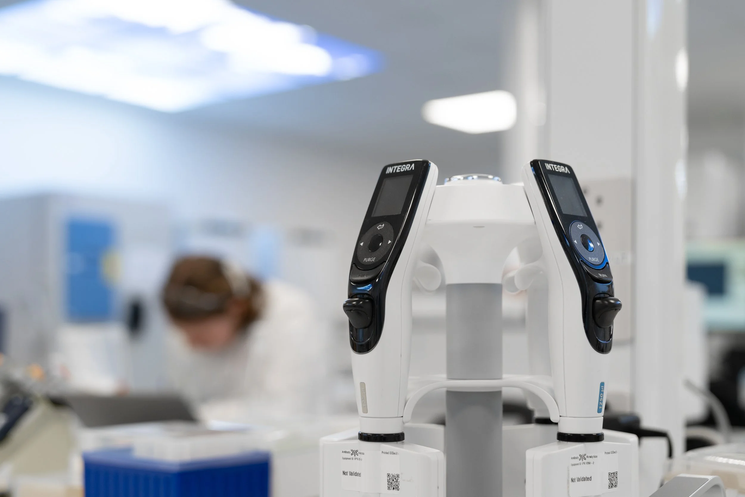 Close-up of electronic pipettes mounted on a stand in a biotech lab, with a scientist working in the background
