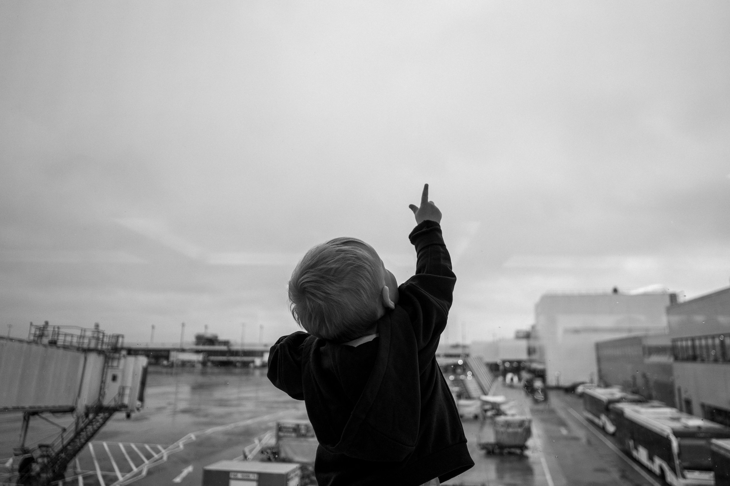 A young child with blonde hair in a black hoodie reaching up and pointing toward the sky at an airport, with planes and airport buildings in the background.