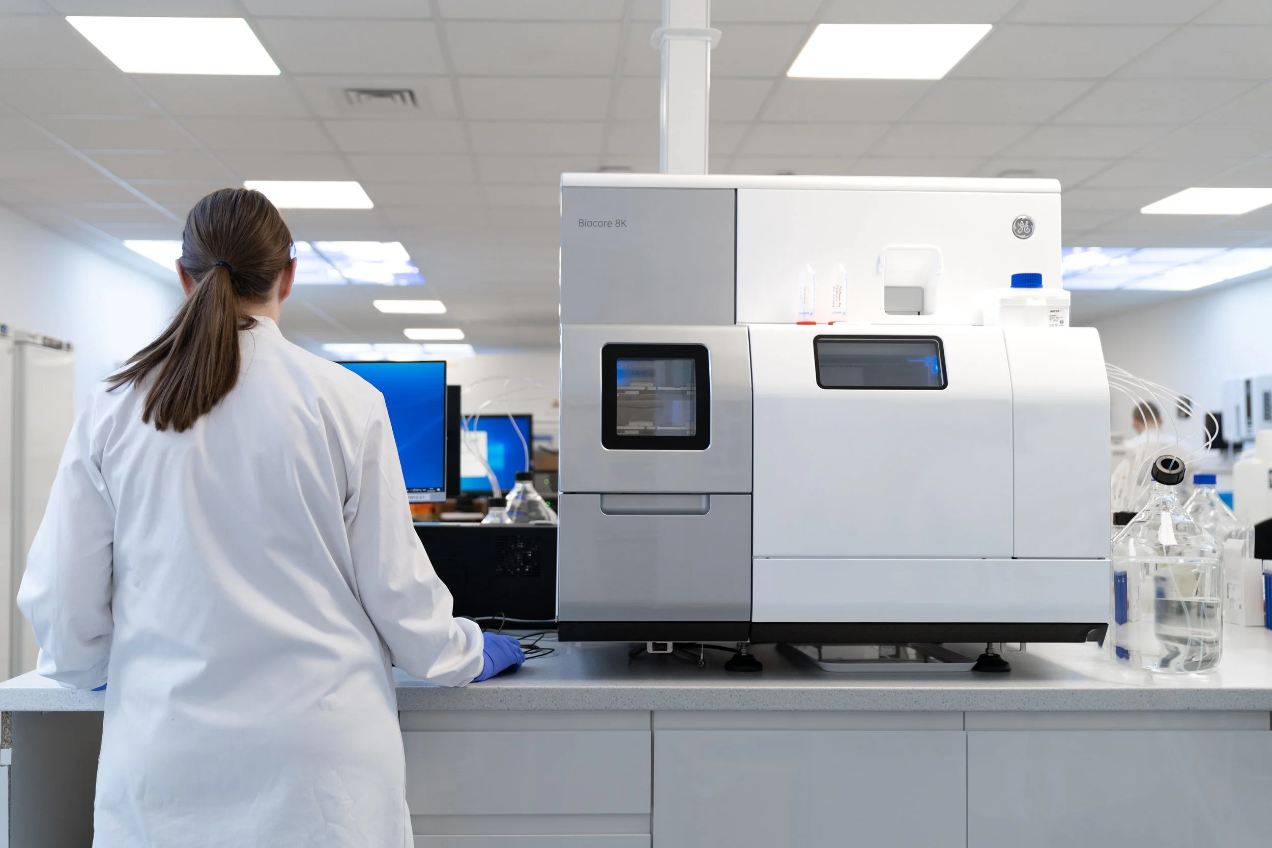 Scientist standing at a Biacore lab instrument, viewed from behind in a biotech research lab