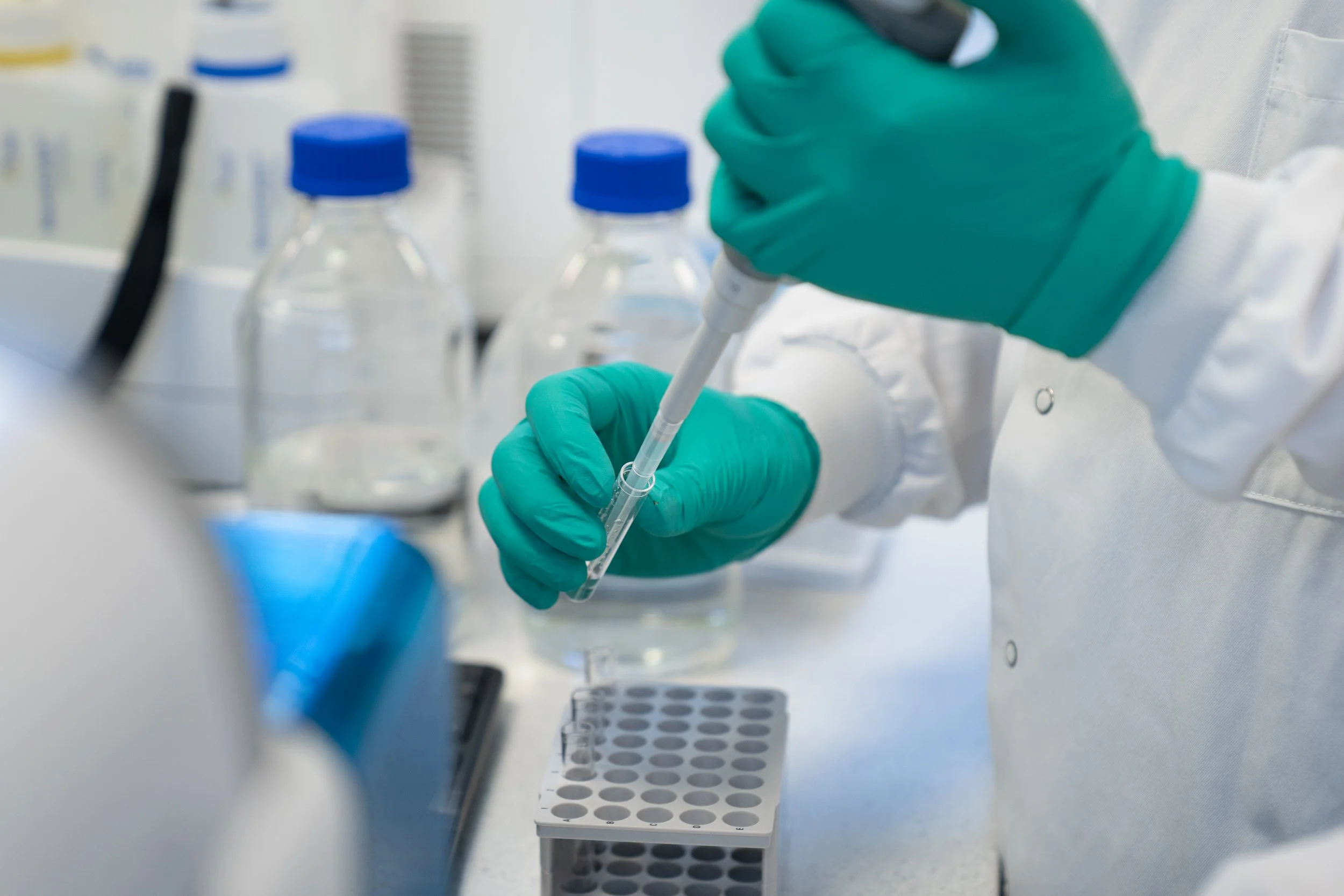 Gloved hands using a pipette to transfer liquid into a sample tube rack during lab testing