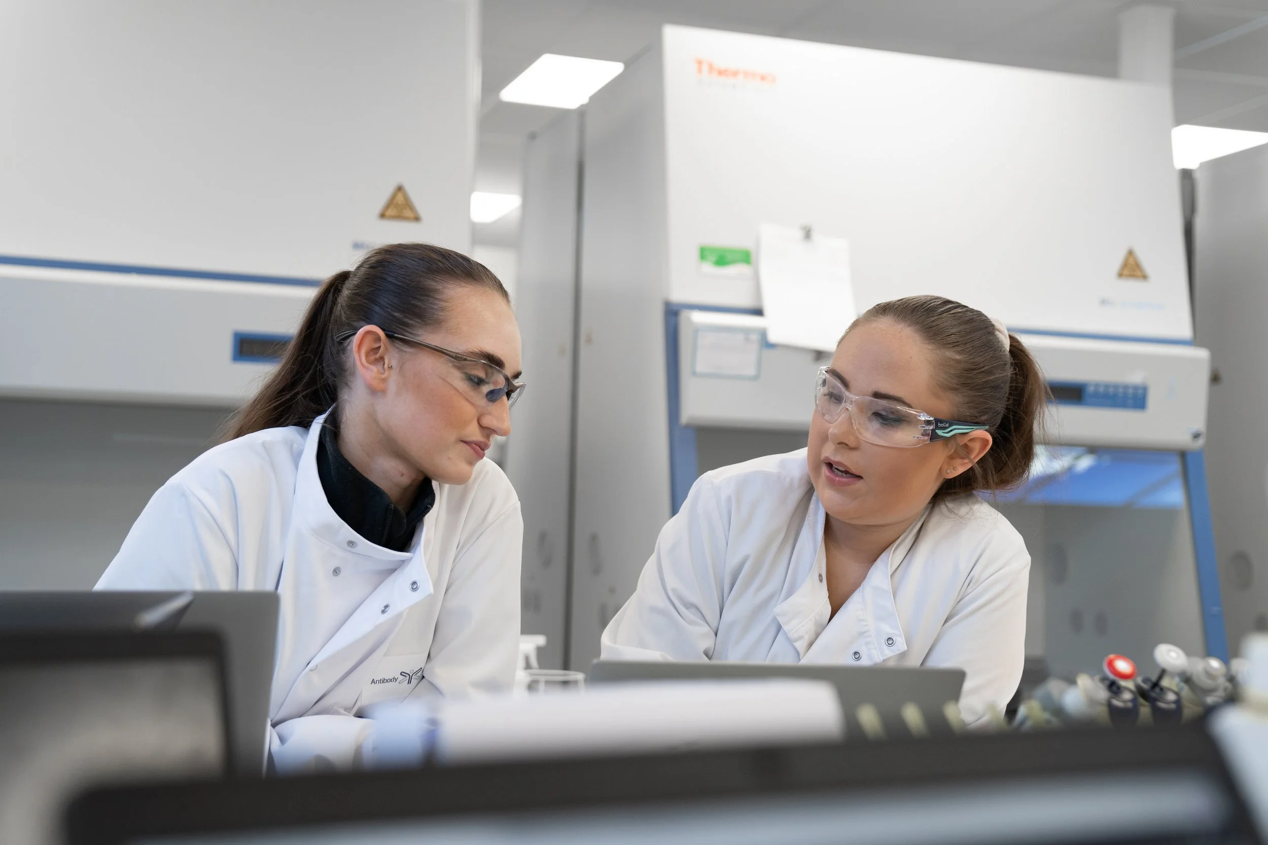 Two scientists in lab coats and safety glasses discussing results at a biotech lab bench