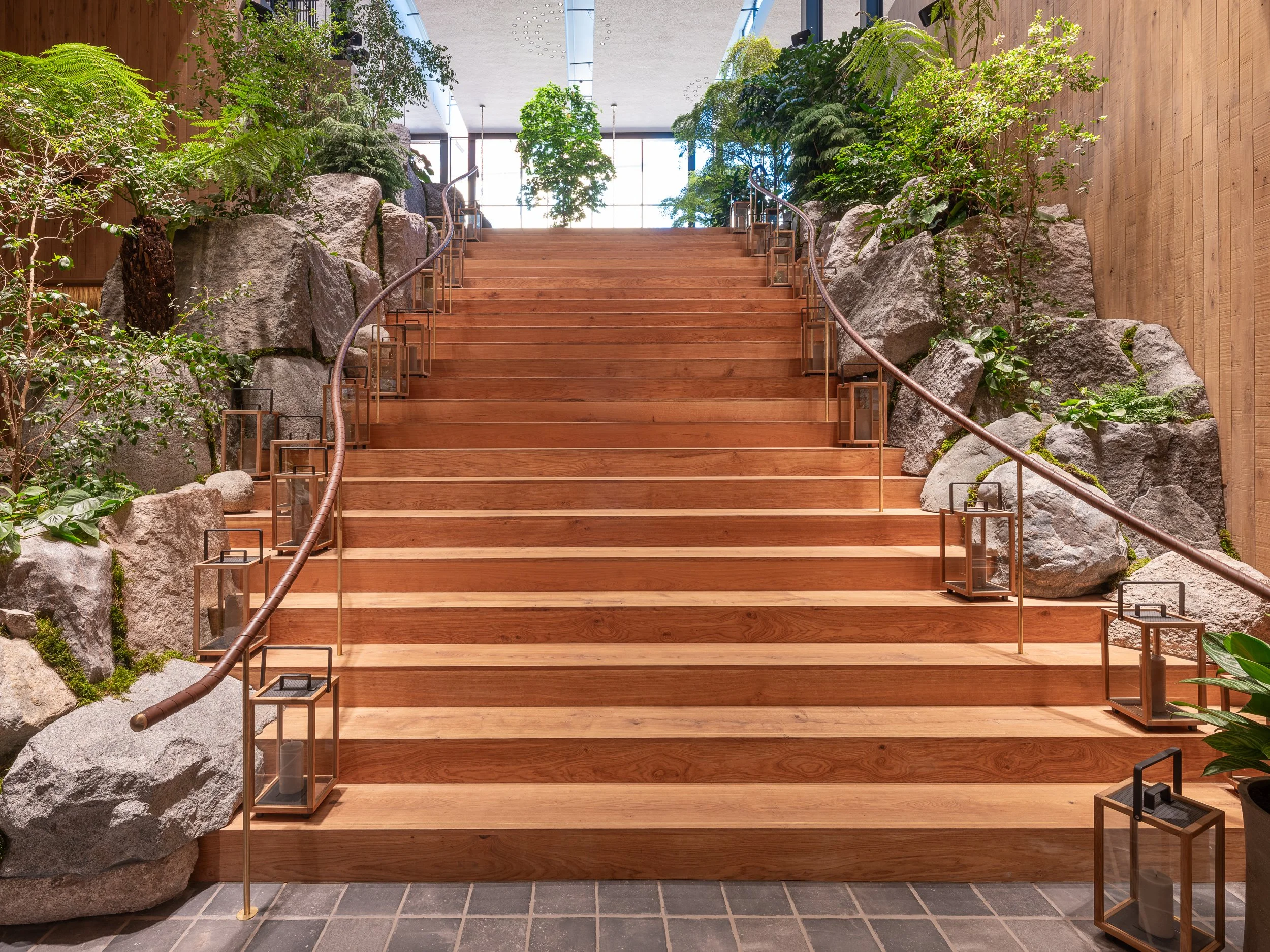Indoor wooden staircase with large rocks and green plants on either side, metal handrails, and lanterns lining the steps, leading up to a large window with natural light.