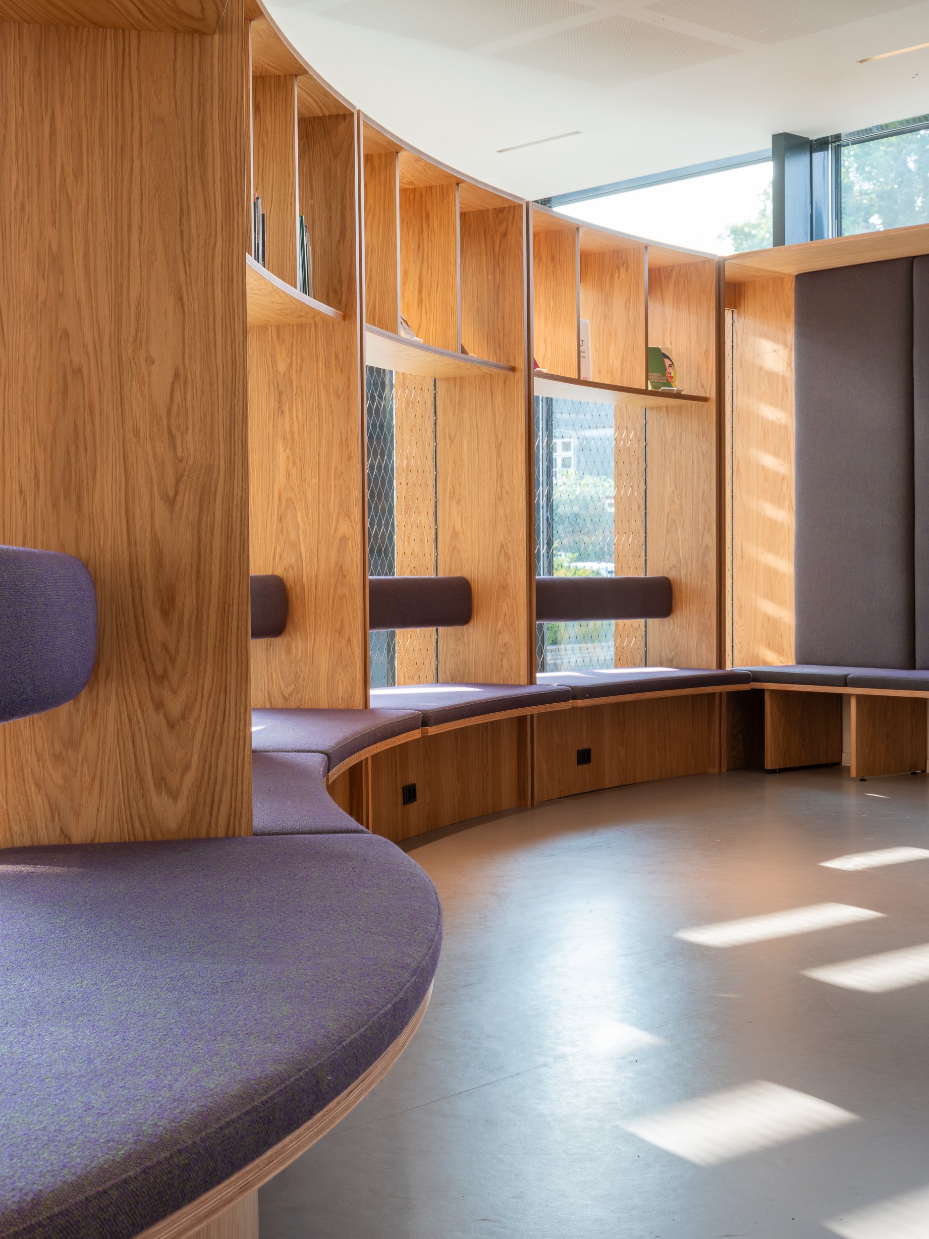 Interior view of a modern space with curved wooden wall and built-in seating, large windows allowing natural light, and a few books on shelves.