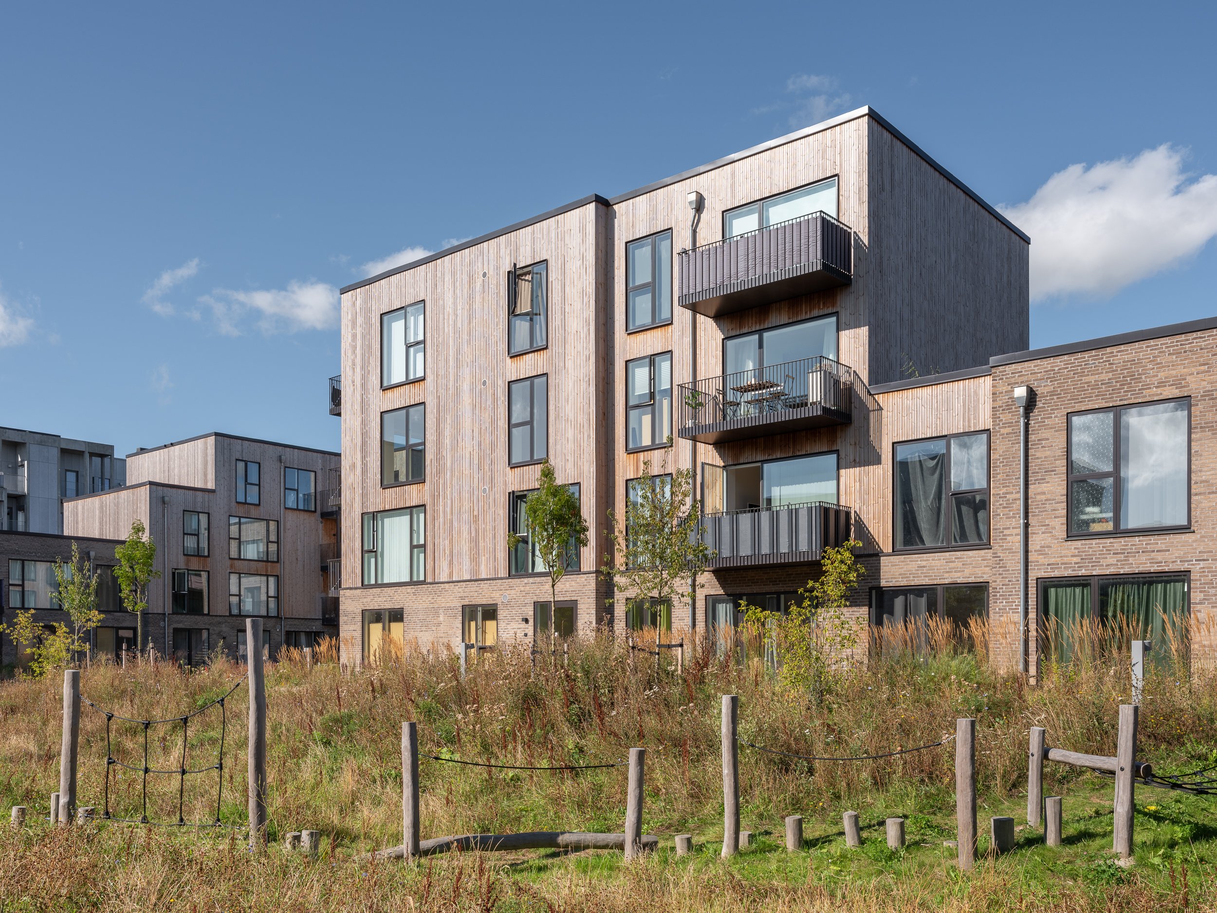 Modern multi-story apartment buildings with wooden and brick facades, several balconies, and large windows, set against a blue sky with a few clouds, with a grassy foreground and small trees.